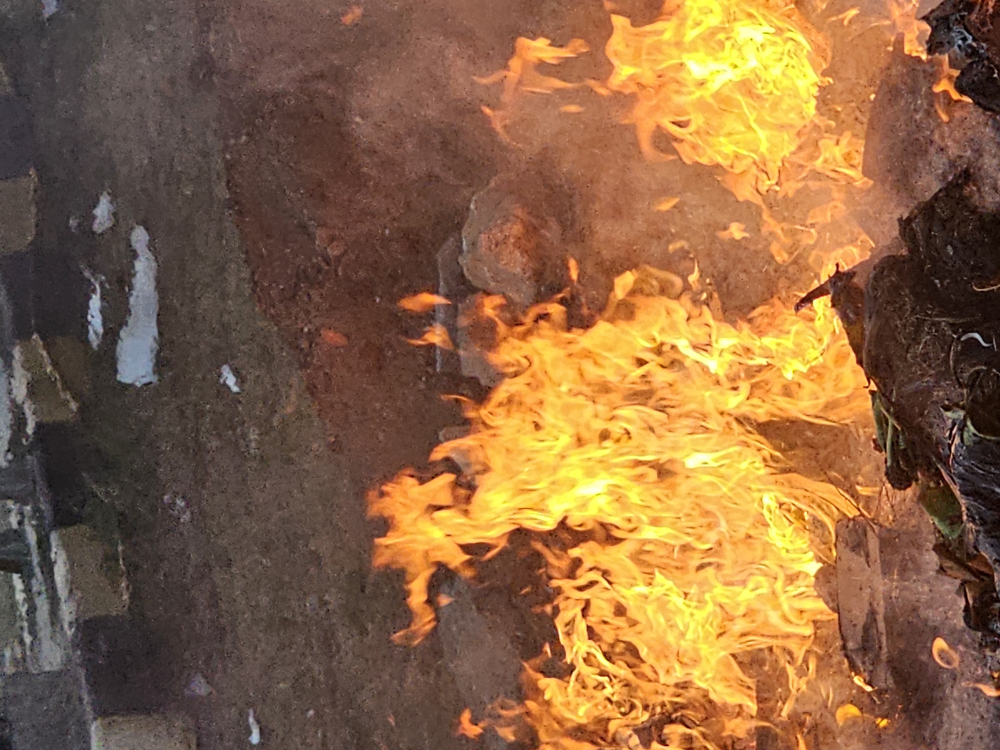 The image captures a close-up view of a fire burning outdoors. The flames are vibrant orange and yellow, with some areas appearing almost white from intense heat. The fire is consuming what appears to be debris, including dark, matted material and some lighter-colored objects that could be rocks or larger pieces of refuse. The background consists of dry, reddish-brown earth, with some lighter patches that might be snow or foam. In the upper portion of the image, there are indistinct shapes that resemble concrete blocks or gravestones, suggesting the fire might be located in a cemetery or an area with such structures. The overall impression is one of uncontrolled burning in an outdoor, possibly rural or undeveloped, setting. The location in Denpasar, Indonesia, is not visually evident from the image itself; this information would need to be provided externally.