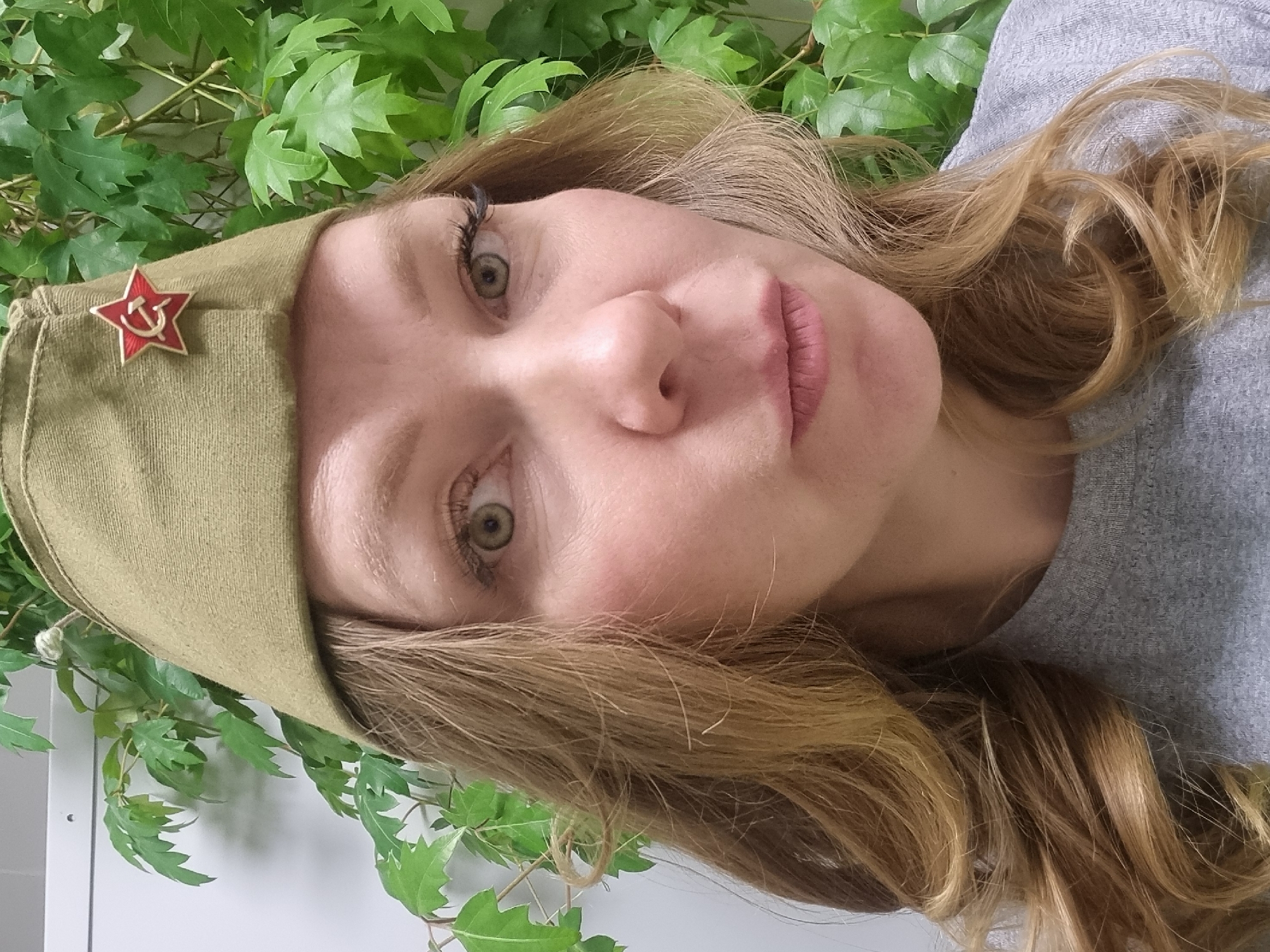 A woman is wearing a light green garrison cap with a red star emblem, superimposed with a gold hammer and sickle. The background includes green leafy plants. The woman is wearing a gray top. The location is not specified, and there is no evidence to support it being Kemul, Russia.