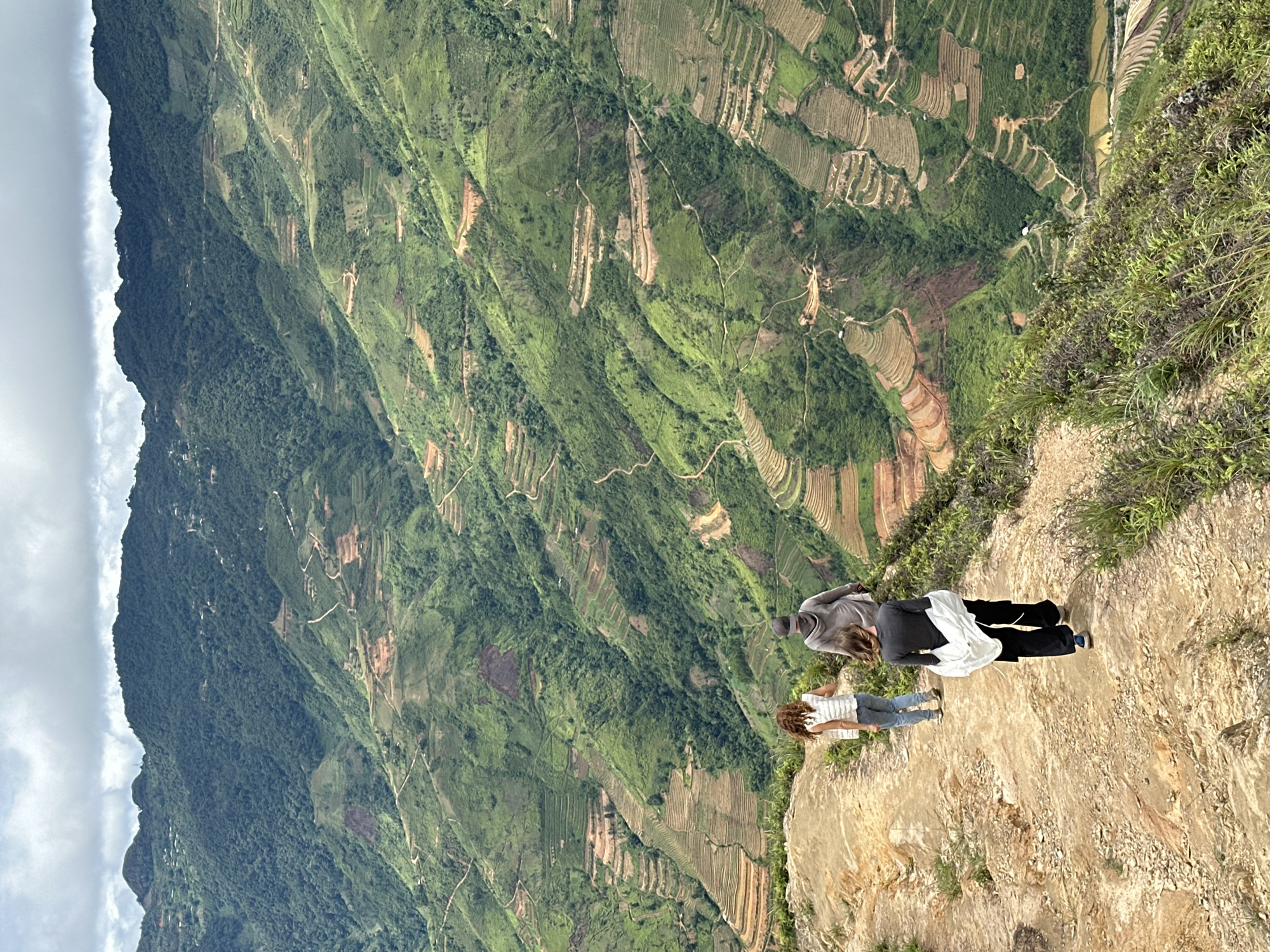 Three people are walking on a dirt path along a mountain ridge with a view of terraced fields in Ta Xua, Vietnam. The person in the foreground, wearing a dark gray long-sleeved shirt and black pants, has a white garment draped over their left arm and is walking away from the camera. Behind them, a person wearing a light gray hooded sweatshirt and a dark cap is walking in the same direction, followed by a person in a white vest and blue jeans. The background features steep, verdant mountains covered in numerous rice terraces, indicative of the agricultural landscape of the region. The sky is overcast with some patches of lighter clouds. The path appears to be made of rocky dirt with sparse green vegetation growing on its edges.
