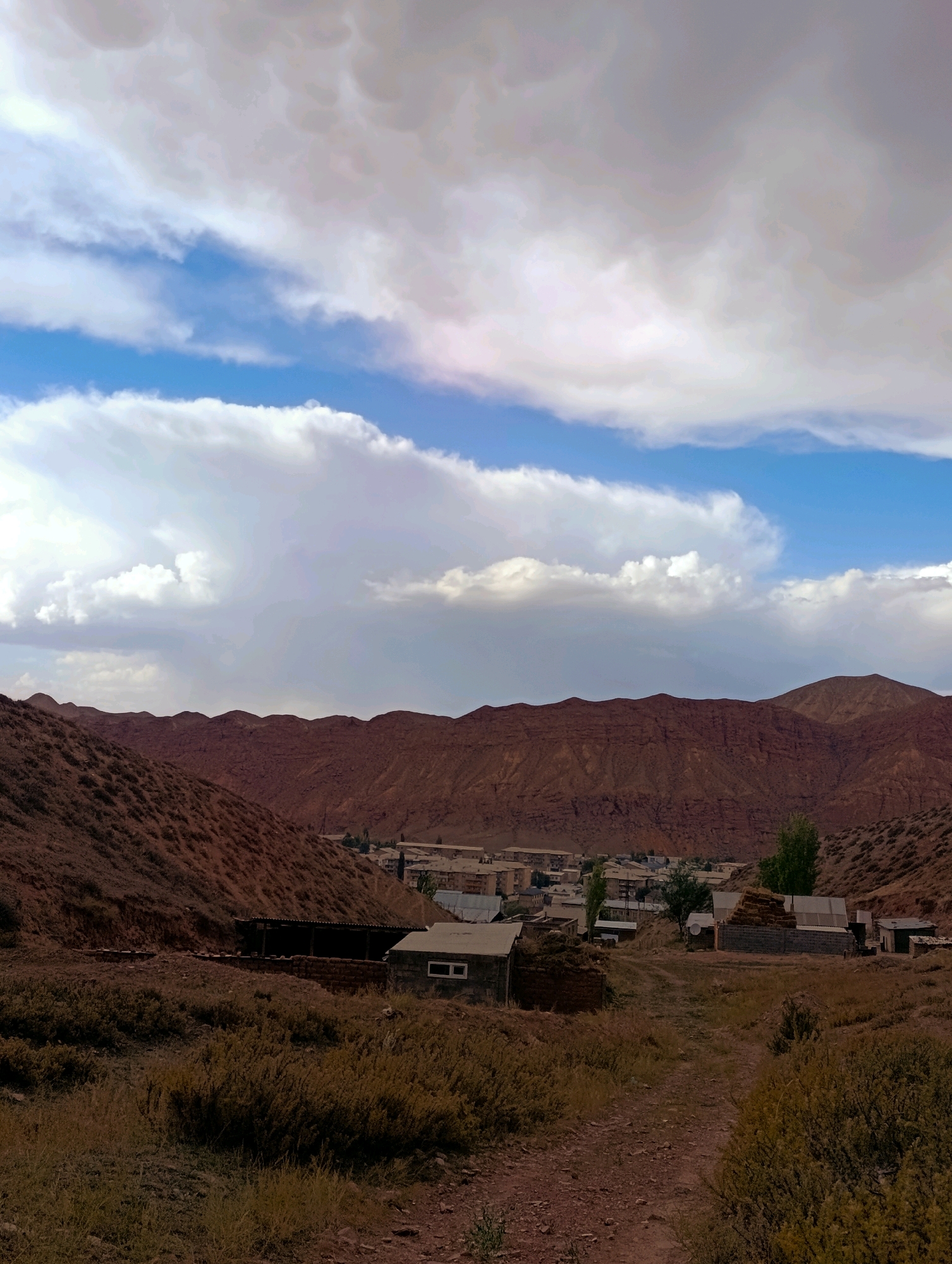 The image depicts a rural landscape in the vicinity of Naryn City, Kyrgyzstan. The foreground is dominated by dry, earthy terrain with sparse vegetation. A dirt path leads into the middle ground, where several rustic buildings and structures are visible. These appear to be homes and agricultural outbuildings, some constructed from brick and others with corrugated metal roofs. One building has a distinctive rectangular window. Behind these structures, a small cluster of more modern buildings, possibly residential apartments or communal buildings, can be seen. The middle ground transitions into the background, which features rolling, reddish-brown mountains. The sky above is dramatic, filled with a mix of blue sky and large, textured clouds, some of which appear to be mammatus clouds, suggesting unsettled weather. The overall scene conveys a sense of a remote, mountainous settlement under a dynamic sky.