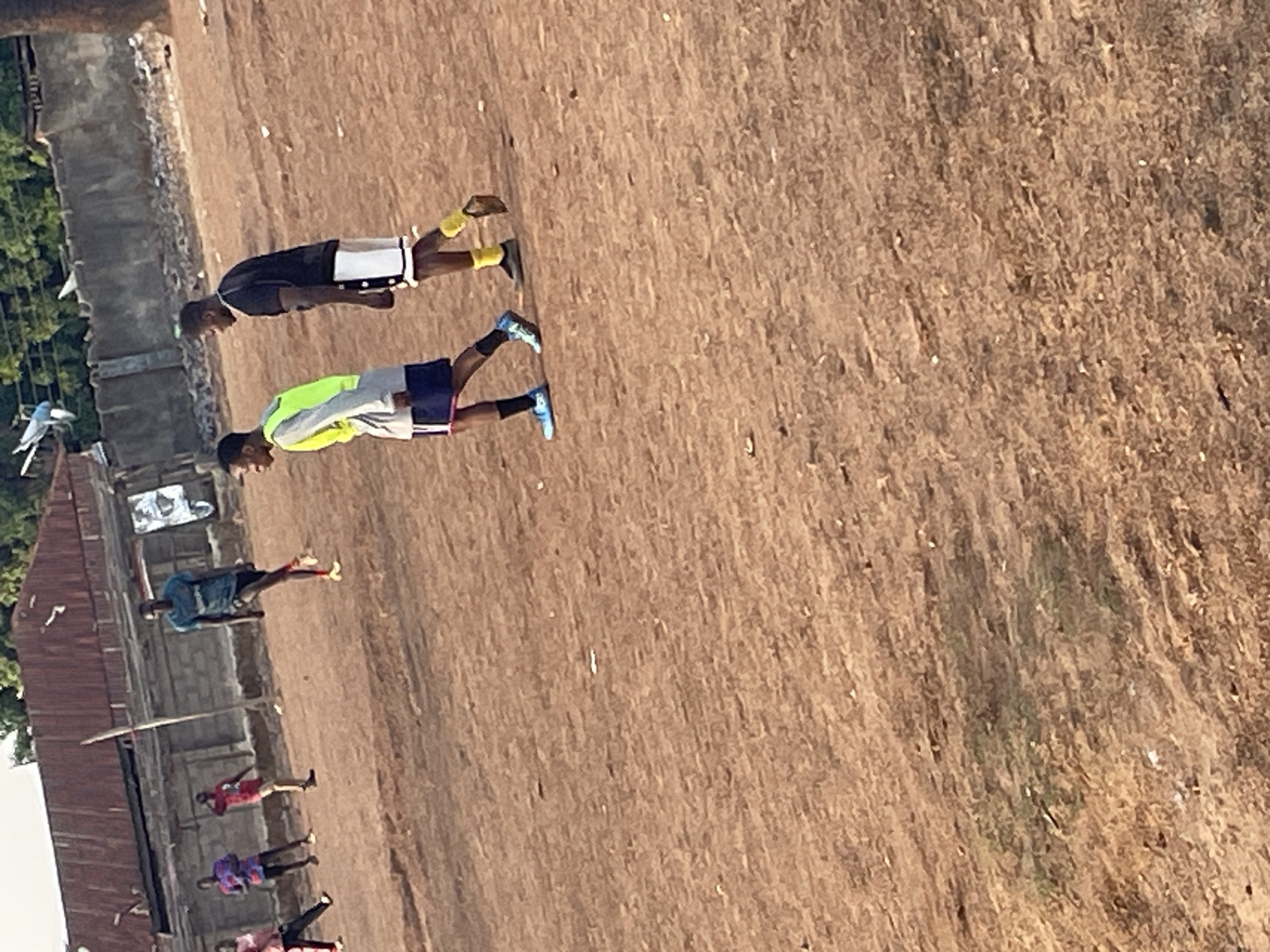 The image depicts an outdoor scene in Akwanga, Nigeria. The environment appears to be a dirt field. Several people are present, some engaged in physical activity. Two men stand out: one wears a bright yellow vest and blue shoes, and the other a black shirt with white shorts and yellow shoes. Other individuals are visible in the background near what looks like a building or structure.