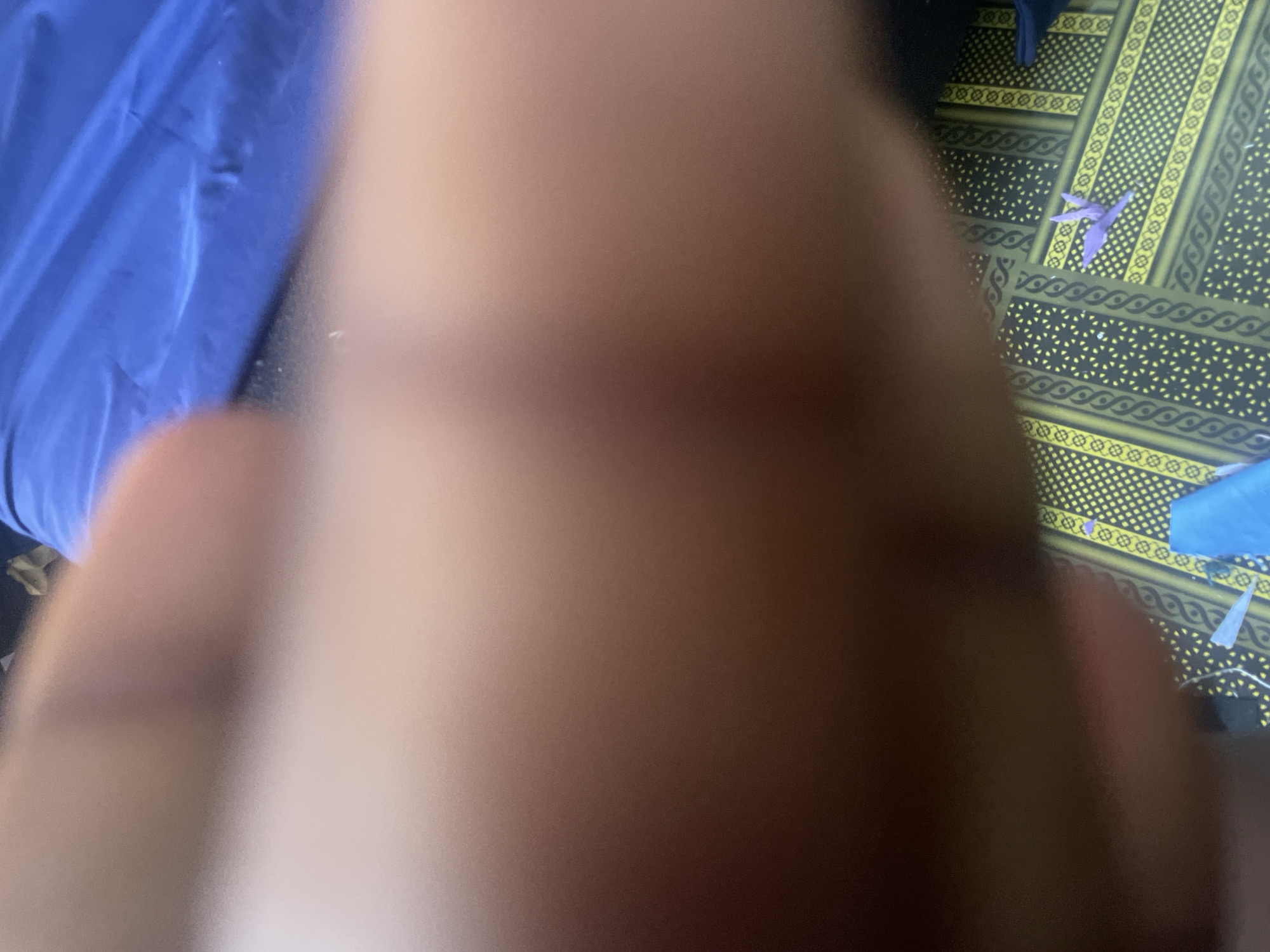 The image shows a close-up view of a person's hand or arm, blurred and out of focus. The hand is positioned over a patterned surface. The pattern appears to be yellow and black, with repeating geometric designs. In the lower right portion of the image, a small purple origami bird is visible on the patterned surface. A blue object, possibly fabric or plastic, is also partially visible on the surface. The background is out of focus but appears to be a dark color, possibly clothing or bedding. There is no indication of the location being Jalingo, Nigeria, as the image lacks any identifiable landmarks or context.