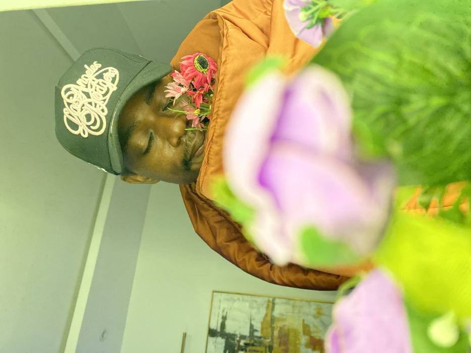 An upside-down close-up reveals a person wearing a black cap with an intricate white design, an orange puffy jacket, and flowers tucked near their face. The person's eyes are closed. Dominating the foreground are blurred purple and green flowers. The background shows a glimpse of a room with a painting on the wall and a light-colored wall. The overall image has a soft, dreamy quality due to the focus and color palette.