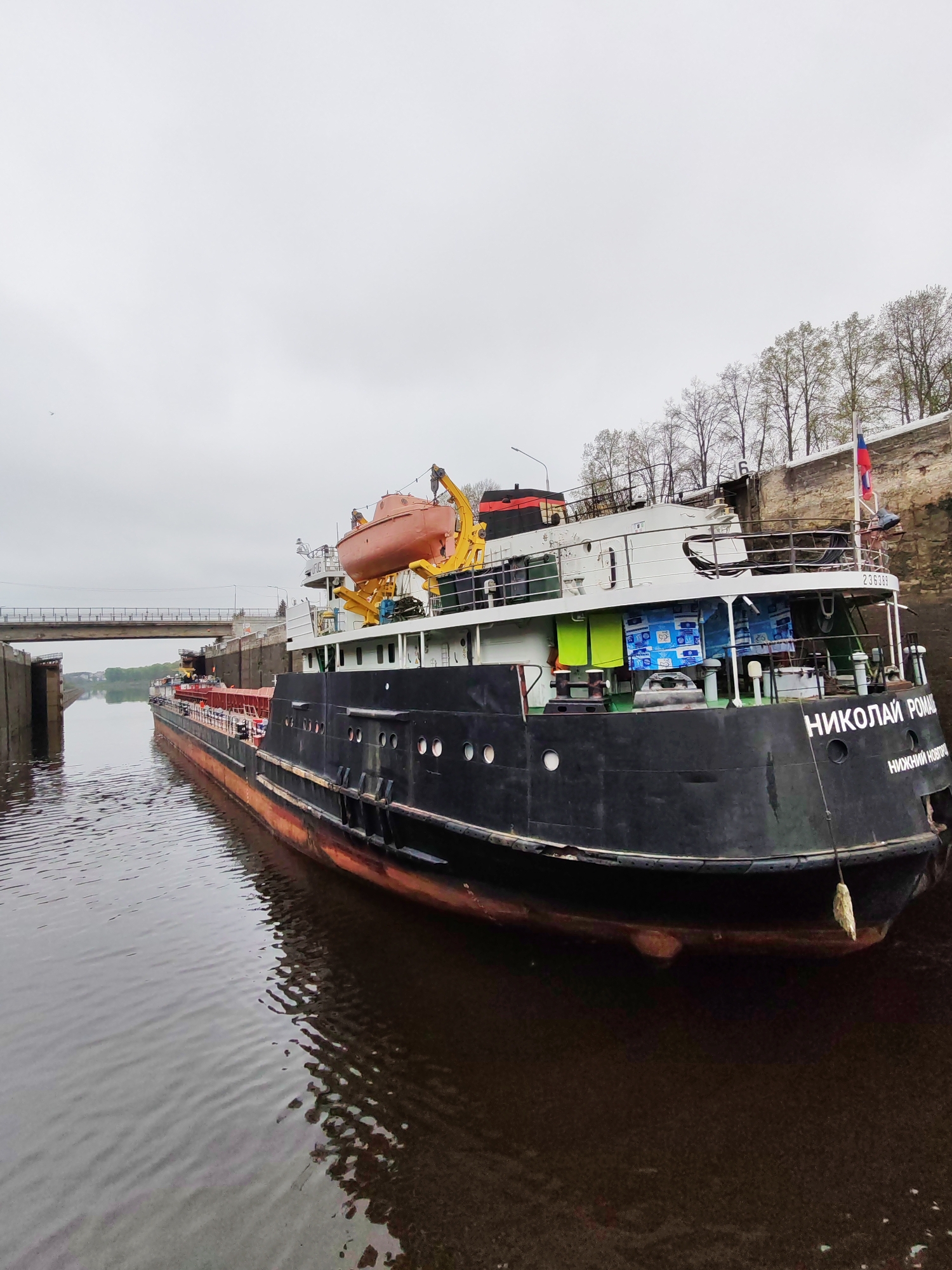 The image depicts a large, black ship, named "НИКОЛАЙ РОМА" (Nikolay Roma), moored in a canal. The ship is docked beside a stone embankment with a few bare trees visible above. A bridge spans the canal in the distance, and the sky is overcast. The water in the canal is dark and reflects the ship's hull. The ship is docked to the embankment and the city of Nizhny Novgorod is printed at the stern of the ship.