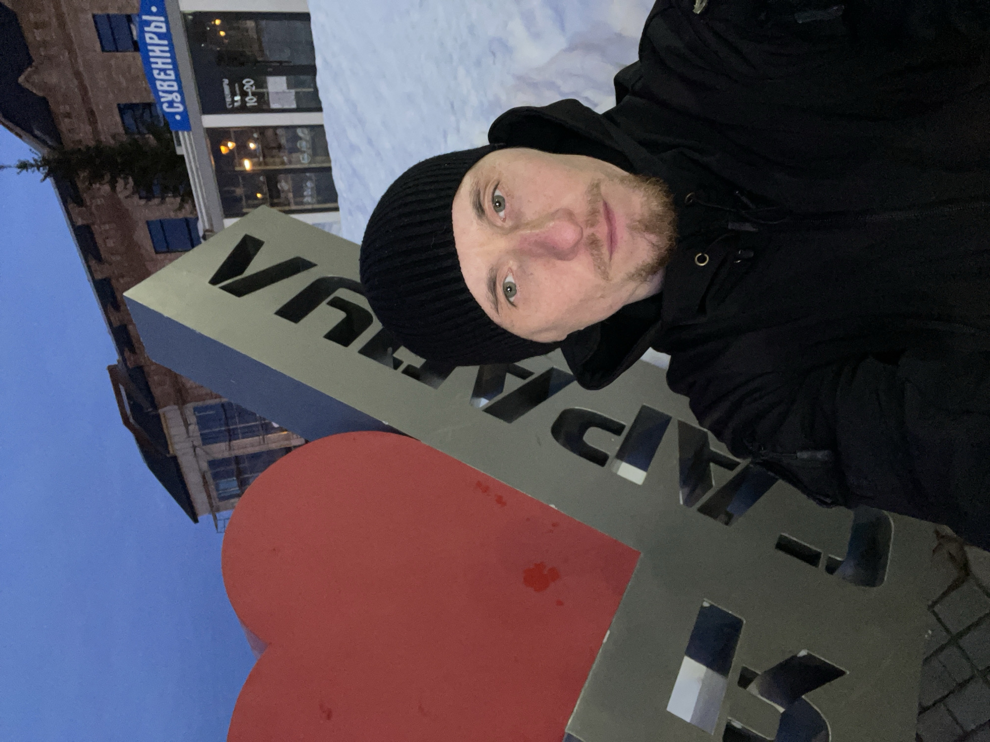 The image displays an outdoor scene in Sarapul, Russia, captured during twilight or dusk, characterized by a deep blue sky. A male individual, appearing to be in his late 20s to early 40s, is positioned in the foreground, looking directly at the camera. He wears a black knit beanie and a black hooded winter jacket. Facial hair consists of a short goatee.
To the subject's left is a large public art installation. This installation features a prominent, partially obscured red heart shape, followed by the Cyrillic word "САРАПУЛ" (Sarapul) constructed from metallic, cut-out letters against a dark grey or silver background. The base of the installation rests on a patterned dark paved surface.
The environment includes snow covering the ground to the right of the subject and the sign. In the background, a two-story brick building under construction or renovation is visible to the left, featuring scaffolding and exposed brickwork. To the right, behind the snow, is a commercial building with a blue sign displaying "СУВЕНИРЫ" (Souvenirs) and an additional sign with text including "10-20," likely indicating business hours. Interior lights are visible through the storefront windows. A conifer tree stands between the background buildings.
