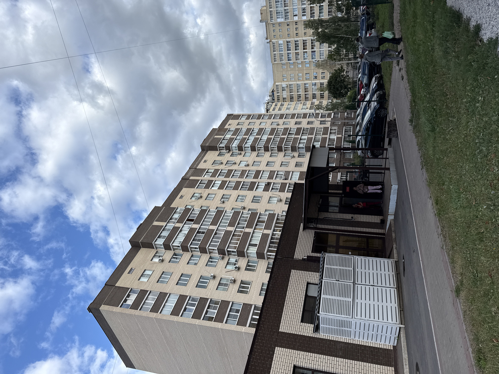 The image depicts a street scene in Zheleznogorsk, Russia, featuring an apartment complex and a smaller brick building. The sky is cloudy with patches of blue. Two individuals are walking away from the viewer on the right side of the frame, one carrying a green plastic bag. Several cars are parked in front of the apartment complex. The apartment building is tall and modern, with many windows. A low brick building is in the foreground, adjacent to the apartment complex. White louvred structures are positioned outside the brick building. The overall environment appears to be an urban residential area.
