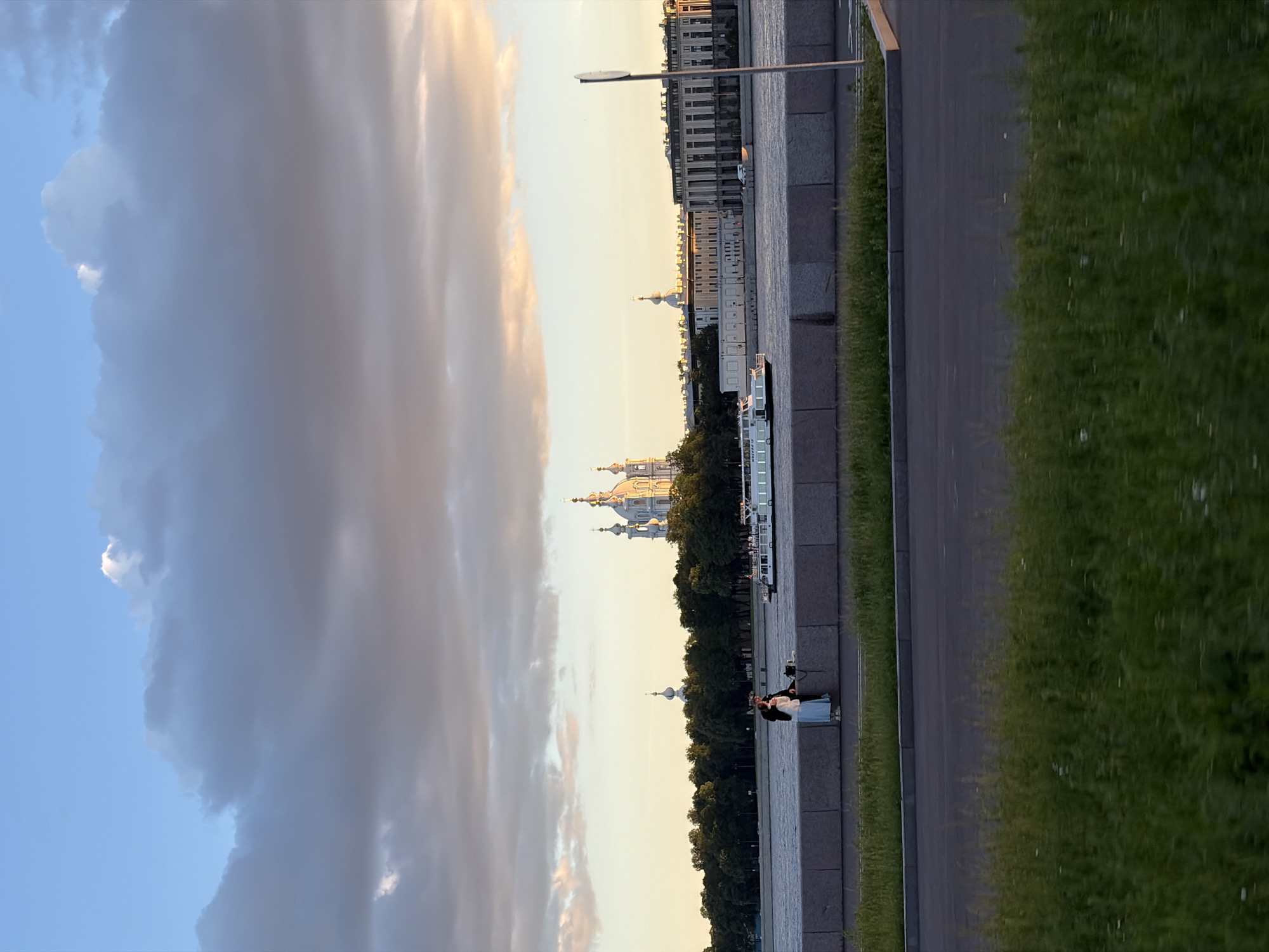 This image depicts a view across a river in Saint Petersburg, Russia, likely the Neva River, towards the Smolny Cathedral complex. The sky is filled with dramatic clouds, with patches of blue sky visible and sunlight illuminating the tops of the clouds, suggesting either sunrise or sunset.
In the foreground, a paved walkway or embankment runs along the river, topped by a granite wall. Two individuals, a man and a woman, are seated on this wall, facing the river. The woman is wearing a light blue dress, and the man is in darker clothing. They appear to be engaged in conversation or simply enjoying the view.
On the opposite bank of the river, a line of lush green trees forms a backdrop. Prominently featured behind the trees is the distinctive white and blue Smolny Cathedral, with its multiple domes. To the right of the cathedral, other buildings of architectural significance are visible, including structures with domes and classical facades. A white passenger boat is navigating the river, moving from left to right in the frame. The overall impression is of a serene urban landscape at a picturesque time of day.
