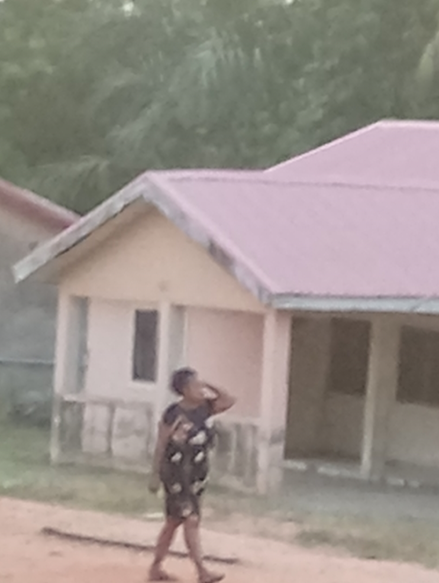 The scene depicts a woman walking along a dirt path in front of a building with a pink roof. The building has a beige exterior and appears to have an open area or porch on one side with visible supports. Trees are visible in the background. The woman is wearing a dark dress with a light-colored pattern and sandals. She has short hair and is holding her hand up near her head. The image has a slightly blurry quality. The location is an unspecified city and region.