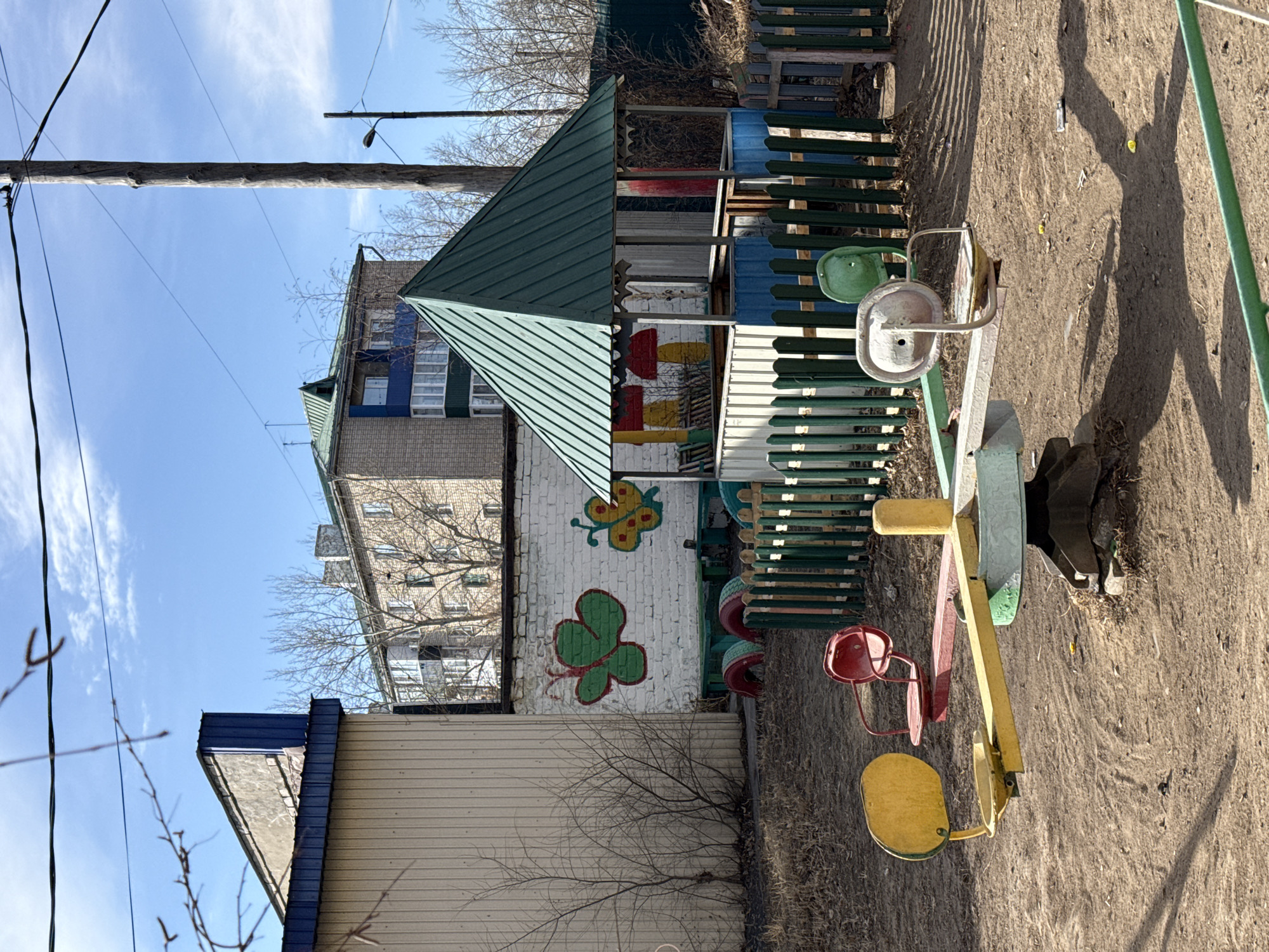 The image captures a playground scene in Borzya, Russia. A child's playground structure with a green roof and painted with images of a butterfly and a four-leaf clover is visible. A seesaw with a yellow and red seat is present in the playground. The ground is dirt. A building is in the background, along with a wooden utility pole and wires against a blue sky with clouds. Bare branches from a small tree are in the foreground. A fence surrounds the playground.