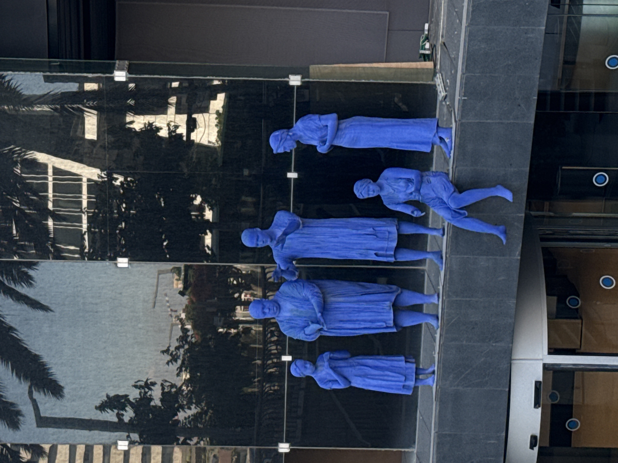 The media file showcases a series of blue sculptures of human figures, likely part of an outdoor art installation. The sculptures appear to be life-sized and are arranged in a line, with some appearing to be walking. They are set against a backdrop of a modern building with large glass panels that reflect the surroundings, including palm trees and other foliage. The glass also reflects the sky, suggesting it is daytime.
The sculptures are monochromatic, rendered entirely in a bright, saturated blue, which contrasts with the darker tones of the building and the reflections. The figures are depicted in a stylized, almost abstract manner, with simplified features and flowing drapery. Their poses suggest movement and perhaps a sense of collective action or procession.
The setting appears to be urban, likely a public space in Tel Aviv, Israel, given the context provided. The architecture of the building, with its dark, reflective surfaces and clean lines, is characteristic of contemporary urban design. The presence of palm trees further reinforces the likely location.
The time of day is not explicitly indicated, but the bright lighting and reflections suggest it is daytime, possibly during sunny weather. There are no discernible emotions on the faces of the sculptures due to their stylized nature. No visible text is present in the image. The overall impression is one of a public art display integrated into the urban environment.