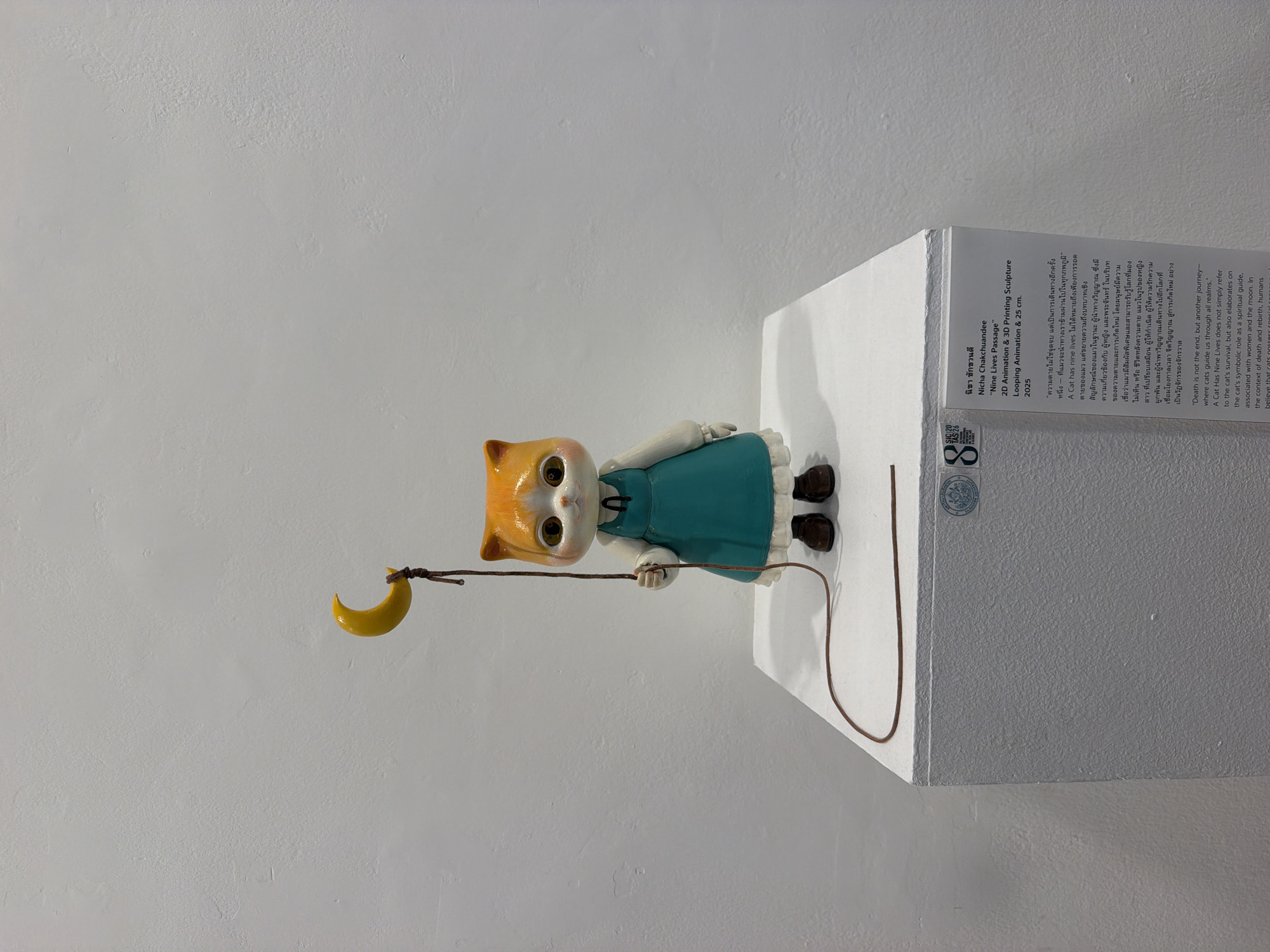 An artwork titled "Nine Lives Passage" by Nicha Chakchuan'dee is displayed on a white pedestal in an indoor exhibition space, likely a gallery in Bangkok, Thailand. The artwork features a doll-like cat figurine with orange and white fur, wearing a teal dress and brown shoes. The cat is holding a yellow crescent moon on a string, which is suspended from above and trails down to the pedestal. The text on a label beside the pedestal provides details about the artwork. The background is a plain, textured white wall.