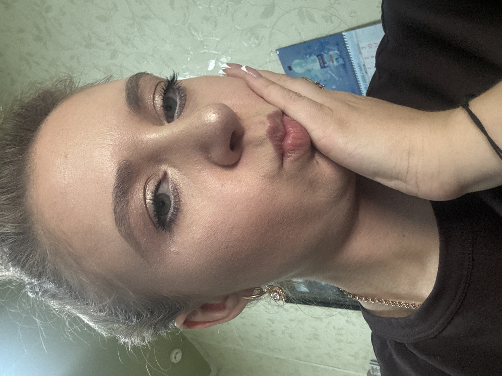 A close-up, self-portrait of a young woman with blonde hair pulled up, wearing makeup and a gold necklace, is shown. She is resting her right hand on her cheek and pursing her lips. She has a gold earring in her left ear. In the background, a calendar showing the month of May is visible. The lighting suggests an indoor setting.