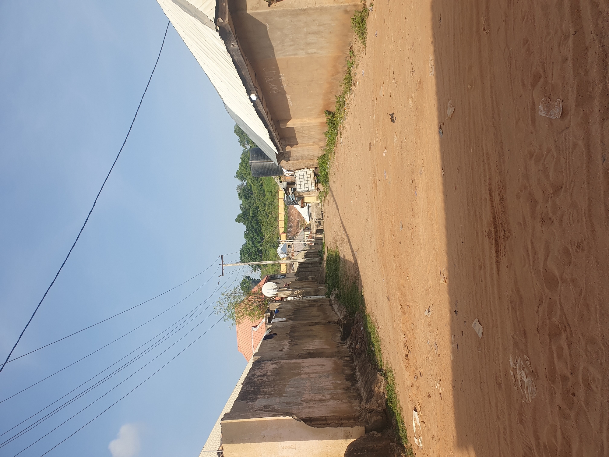 The image depicts a dusty, unpaved road in what appears to be a residential area in Lokoja, Nigeria. On the left side of the road is a tall, weathered concrete wall, and on the right are buildings with corrugated metal roofs. Power lines crisscross the clear blue sky overhead. The road is littered with debris, including plastic bags and other trash. In the distance, more buildings and lush green trees are visible on a gentle slope. The scene suggests a typical urban or semi-urban environment in Nigeria, characterized by its unpaved roads, simple housing structures, and overhead power infrastructure. There are no people visible in the image.