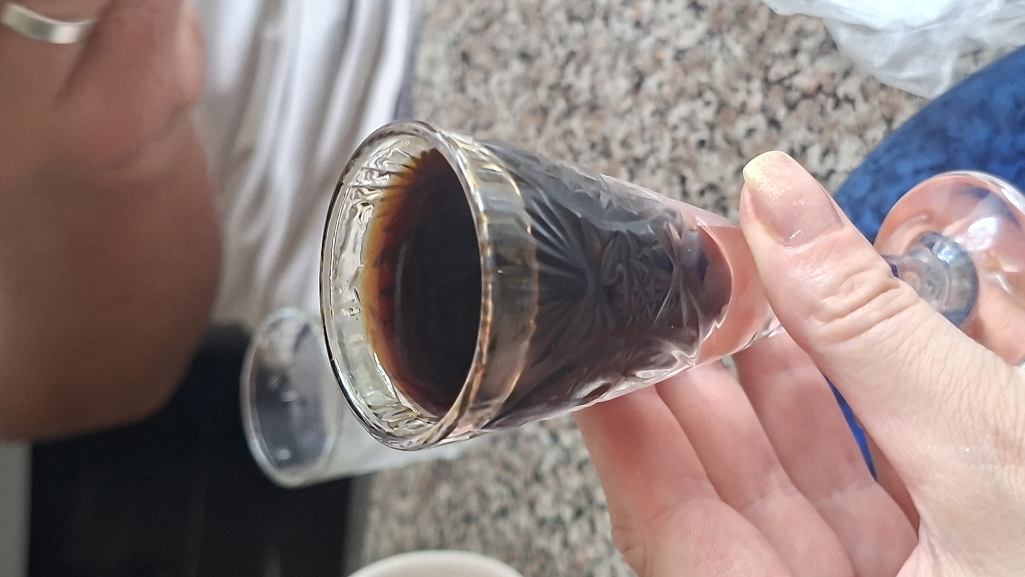 In Myadzyel, Belarus, a close-up shot focuses on a person's hand holding a small, ornate glass filled with a dark liquid, likely an alcoholic beverage or strong coffee. The glass has intricate etched patterns and a light pink liquid at the bottom. In the background, slightly out of focus, a person in a white t-shirt is visible, and another glass of clear liquid sits on a granite-patterned surface. The lighting suggests an indoor setting, possibly a cafe or dining area.