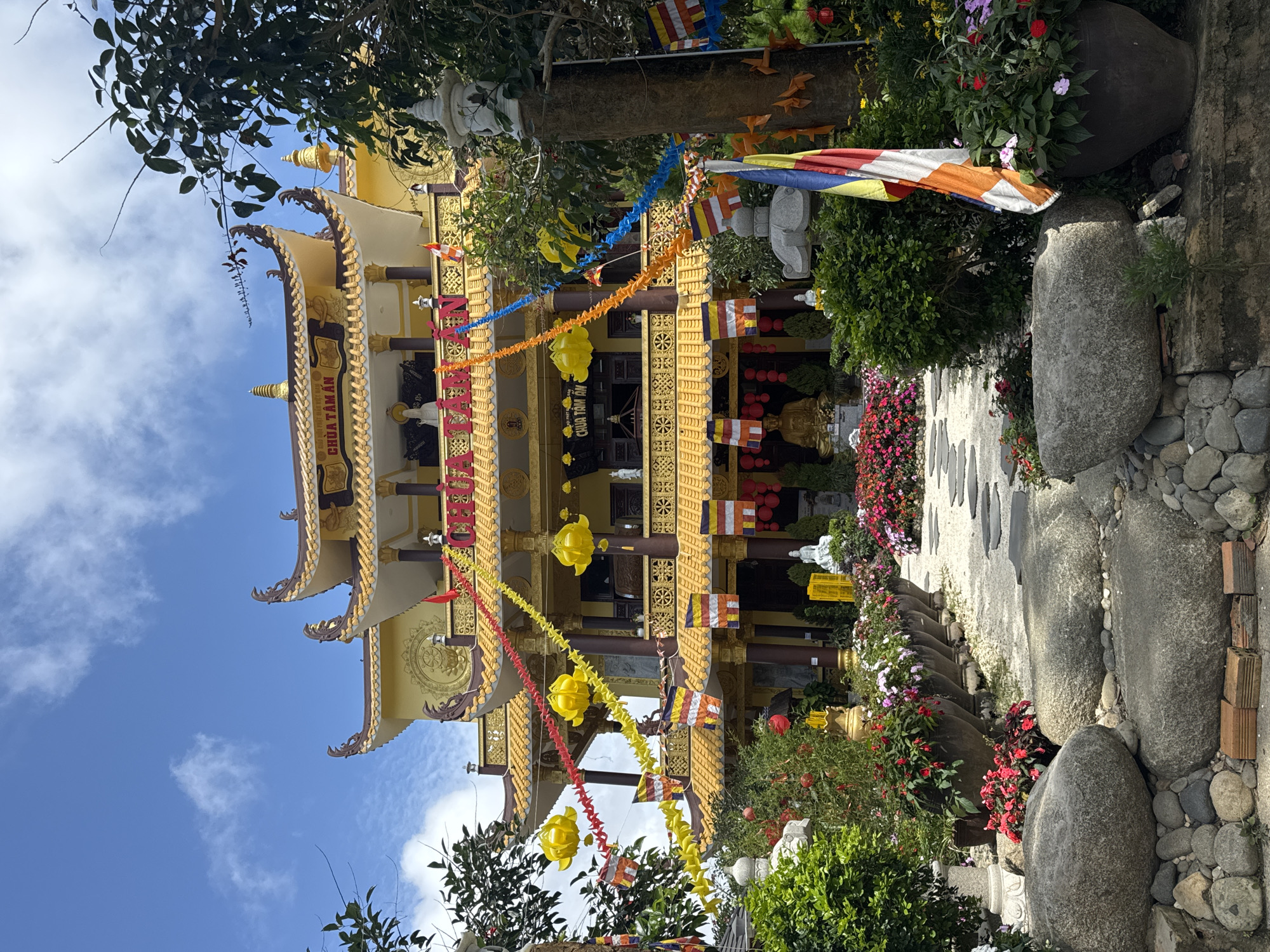 A Buddhist temple, identified by the signage as Chua Tam An, stands prominently in the city of Da Lat, Vietnam. The temple is characterized by its ornate yellow architecture, with multiple tiers of curved roofs adorned with intricate carvings and finials. Red and yellow banners, along with paper lanterns and Buddhist prayer flags, are strung across the facade, creating a festive and colorful atmosphere. In front of the temple, a pathway of stepping stones leads through a garden filled with lush greenery and vibrant flowers in large pots. Large, weathered boulders are placed in the foreground, and several statues, including a seated golden Buddha and white Buddhist figures, are visible within the temple grounds. The sky above is a bright blue, dotted with scattered white clouds, indicating fair weather. The scene captures a serene and spiritual setting amidst a well-maintained garden.