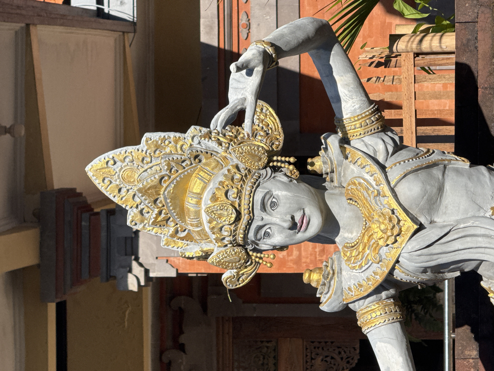 A sideways, medium-shot photograph shows a grey and gold Hindu statue of a Balinese dancer with its left arm raised above its head and right arm and leg bent forward. The statue is in the city of Kutuh, Indonesia.
The statue’s skin is grey, and it is wearing an elaborate gold headdress, shoulder plate, and bracelets. The dancer’s eyes are pale and outlined with a dark, bold line, the nose is broad, and the lips are lightly shaded pink.
The background includes a light-beige building with a tiered, grey exterior and a red building with wood elements. A palm frond and green plant are visible behind the dancer's right arm. The entire scene is well-lit and in focus.