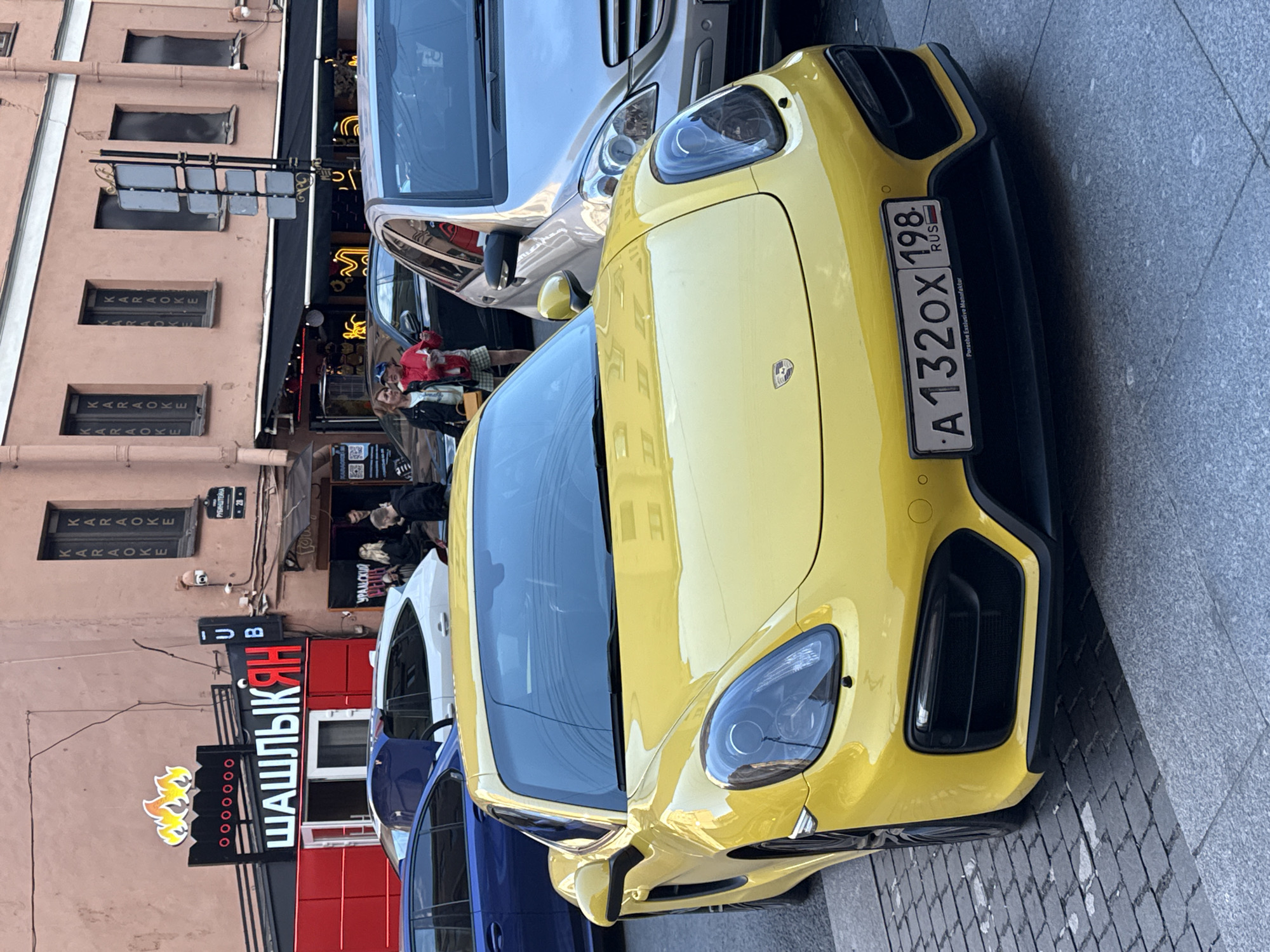 A yellow Porsche sports car is parked on a cobblestone street in front of a building with a sign for "ШАШЛЫК-ЯН" (Shashlik-Yan) which appears to be a restaurant, with "KARAOKE" signs visible on the building's windows. The car's license plate reads "A 132OX 198 RUS". Several people are visible in the background near the entrance of the building and the adjacent establishments. The street appears to be an urban environment in Saint Petersburg, Russia.