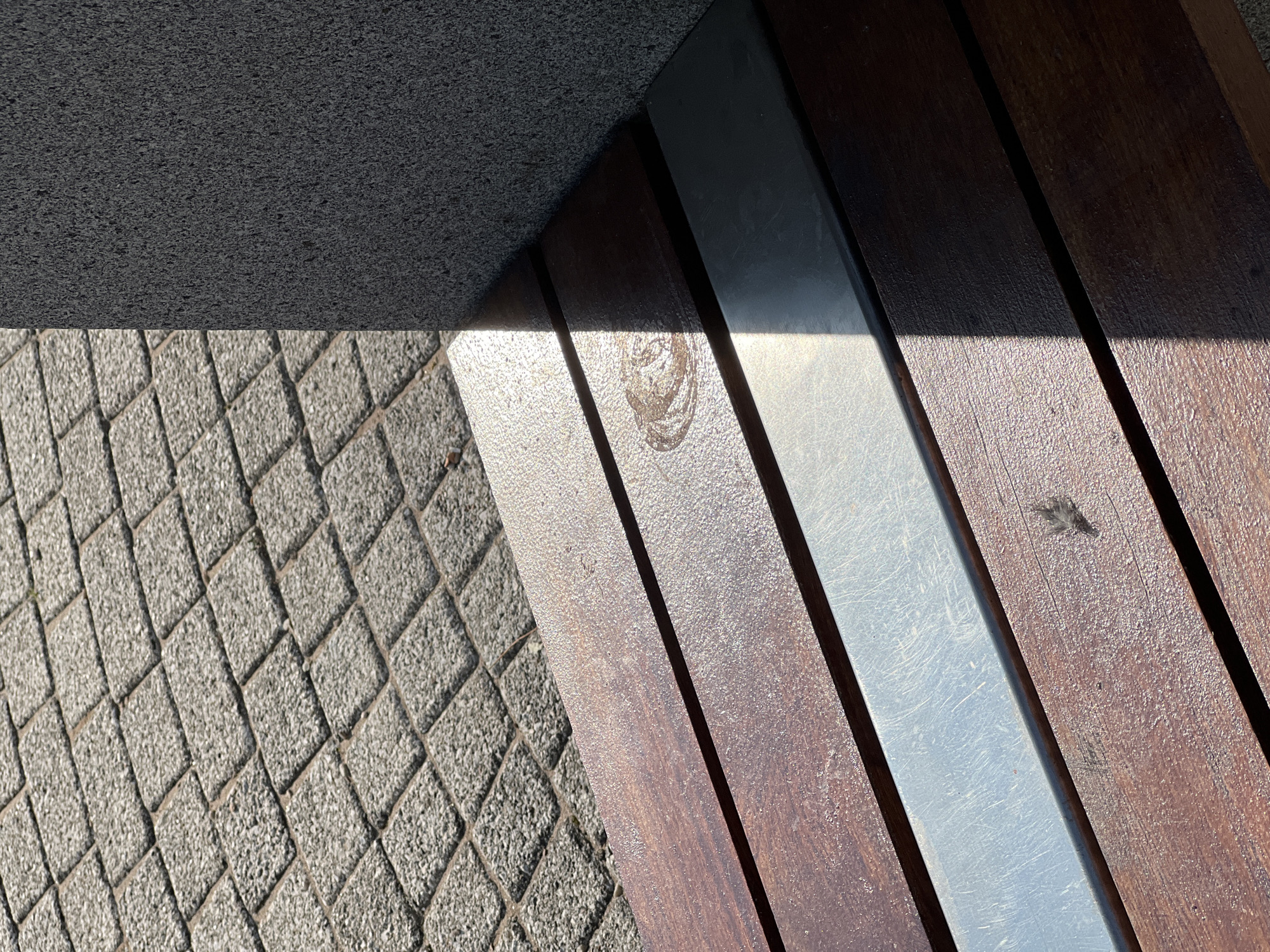 The image shows a close-up of a wooden and metal bench in Donegal, Ireland. The bench is positioned next to a textured, dark grey wall. In the background, there is a paved area with diamond-shaped grey stones. Sunlight casts a sharp shadow across the bench. The bench itself has dark brown wooden slats with a metallic silver strip running horizontally between them. There is a circular imprint on one of the wooden slats, and a small dark object, possibly a feather or debris, is visible on another slat. The overall scene is outdoors, suggesting a public space like a park or walkway.