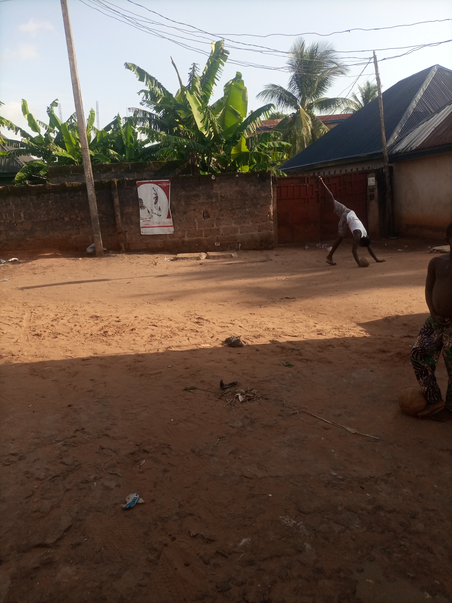 In Nkpor, Nigeria, a dirt road is illuminated by daylight. A person in a white t-shirt and patterned shorts is in the process of a handstand, with their feet in the air and their hands on the ground, near a ball. Another person, partially visible, is standing to the right with patterned pants and appears to be holding a ball. Behind them, a brick wall with a poster attached stands against a backdrop of lush banana trees and palm trees, with corrugated metal roofs of buildings visible above. A utility pole with wires stretches across the sky. The ground is uneven and sandy, with some scattered debris.
