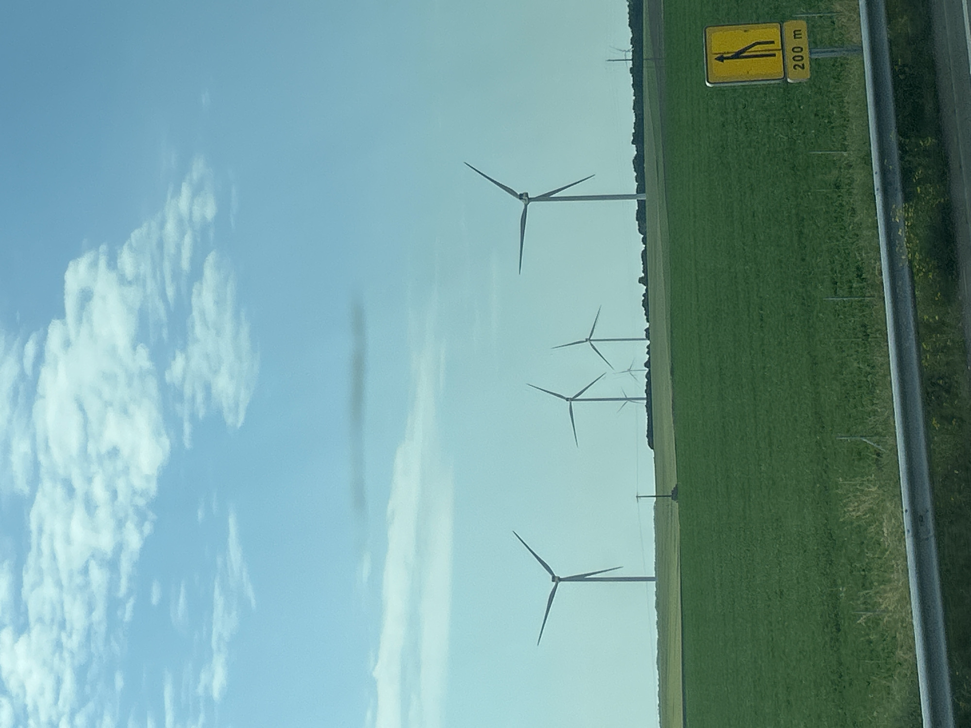 A photograph taken from a moving vehicle shows a rural landscape under a partly cloudy blue sky. In the foreground, a grey metal guardrail runs horizontally across the bottom of the frame. Beyond the guardrail, a vibrant green field stretches into the distance. Several wind turbines are visible in the middle ground and background, silhouetted against the sky. Their blades are varied in their positions, suggesting movement. A yellow traffic sign with black markings indicating a lane reduction is visible on the right side of the frame, with the text "200 m" below it. The environment appears to be an open field or farmland, likely in or near the city of Fontaine-Notre-Dame, France.
