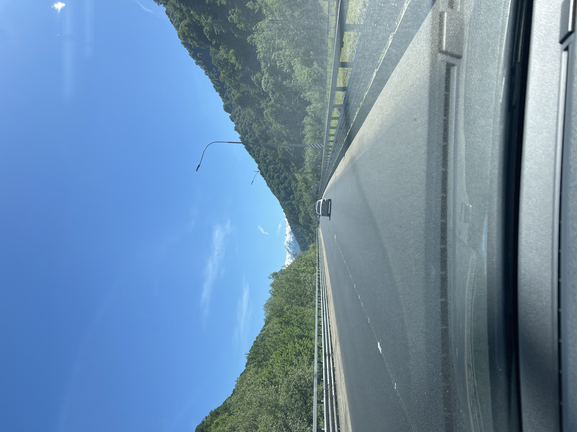 The image captures a bright, sunny daytime scene from inside a moving vehicle on a multi-lane highway, looking forward. The asphalt road, marked with white lines, extends into the distance, with a white car visible ahead in the right lane. A metal guardrail runs along the right edge of the road.
Steep, densely forested mountains and hills rise on both sides of the highway, covered in lush green trees. On the left, a tall street light pole with an extending arm is visible. In the far distance, beyond the immediate green peaks, a prominent snow-capped mountain range is visible under a clear, vibrant blue sky. A few wispy white clouds are scattered across the sky, with bright sunlight creating a slight glare or reflection near the top left corner.
The perspective is from within a car, evidenced by the visible right A-pillar and part of the interior door frame or dashboard on the far right. Some dust or specks are visible on the windshield, indicating it's an on-the-road shot. The overall scene suggests travel through a picturesque, mountainous landscape, consistent with the Chvizhepse region in Russia. No discernible text is visible in the image.