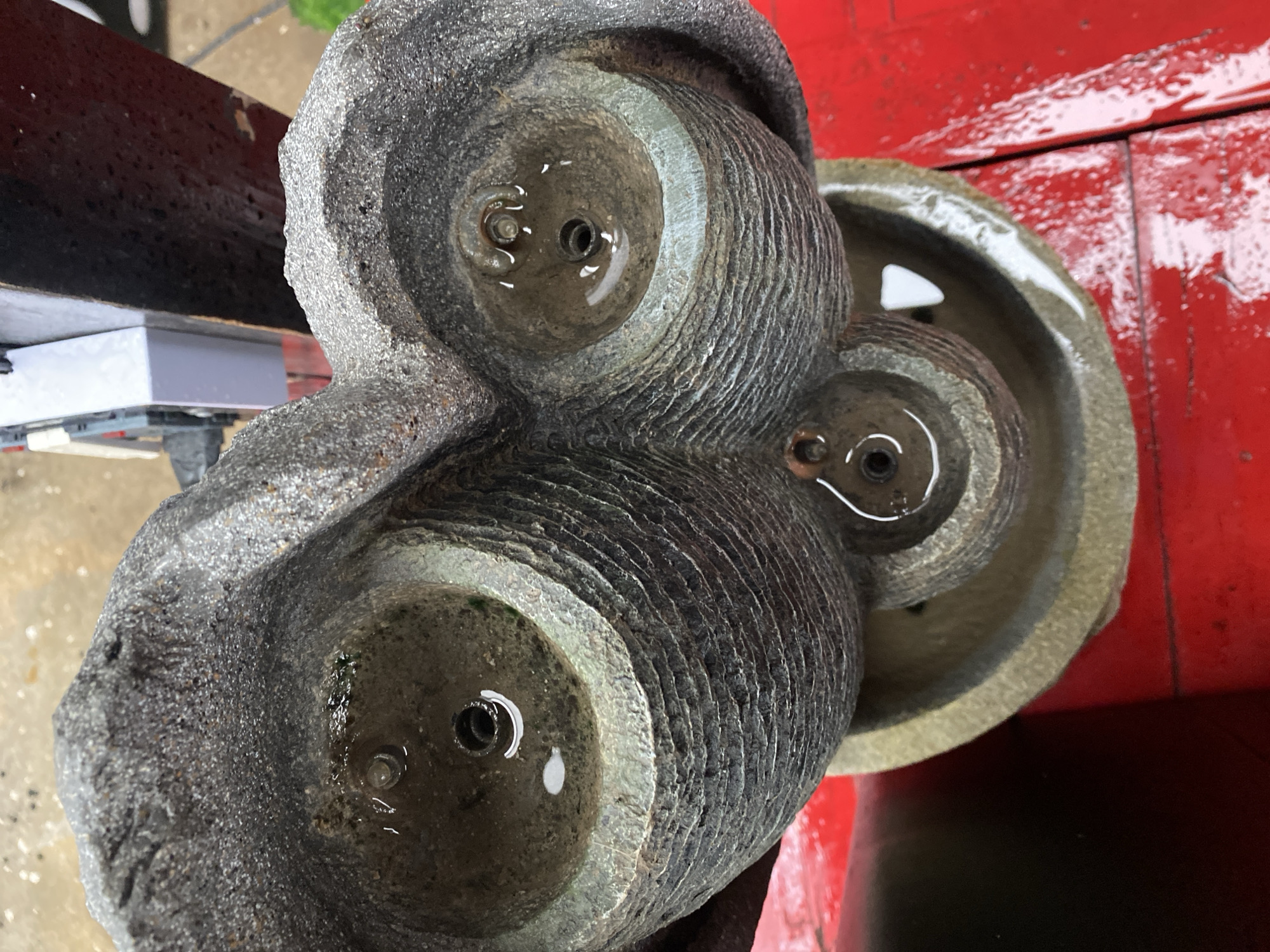A top-down, close-up view of a three-tiered water fountain made of rough, gray stone. Each tier has a circular opening at the top, where water is pooling and trickling down. The stone has a textured, layered appearance with horizontal ridges. The fountain is placed on a bright red surface, which appears to be a table or deck, and is wet from the water. In the background, a white and gray object, possibly an electrical box or appliance, is partially visible. The overall scene suggests an outdoor setting, likely a garden or patio, possibly in the city of Howayi, Nigeria.