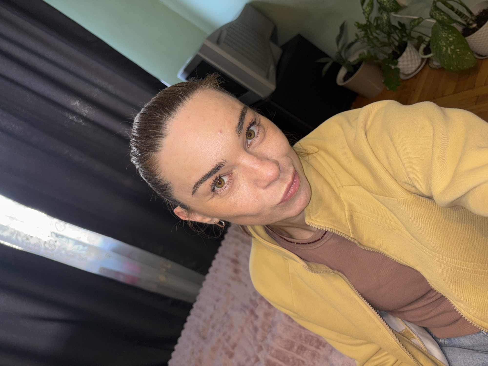 A close-up, selfie-style photograph captures a woman with brown hair pulled back into a ponytail, looking directly at the camera. She has brown eyes and is wearing a yellow zipped jacket over a taupe top. Her face is illuminated by a light source to her left, casting some shadows. The background is out of focus, but appears to be an indoor setting with dark curtains to the left and a pale green wall to the right. Several potted plants are visible on the right side of the frame, along with a portion of a computer monitor. The overall impression is a casual, domestic scene. The location is unknown.