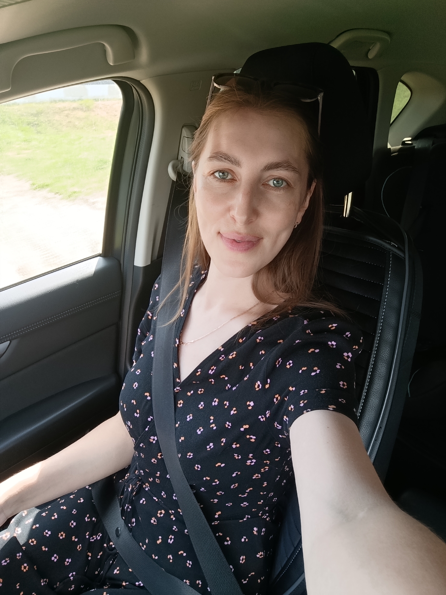 A woman with shoulder-length brown hair and green eyes is seated in the passenger seat of a car, facing the camera and taking a selfie. She is wearing a black dress with a floral print and a seatbelt. The interior of the car, including the black leather seat and the gray door panel, is visible. Through the window, a blurry green landscape with some buildings can be seen, suggesting a location outside a city, potentially in the outskirts of Chaikovsky, Russia. The lighting suggests it is daytime.