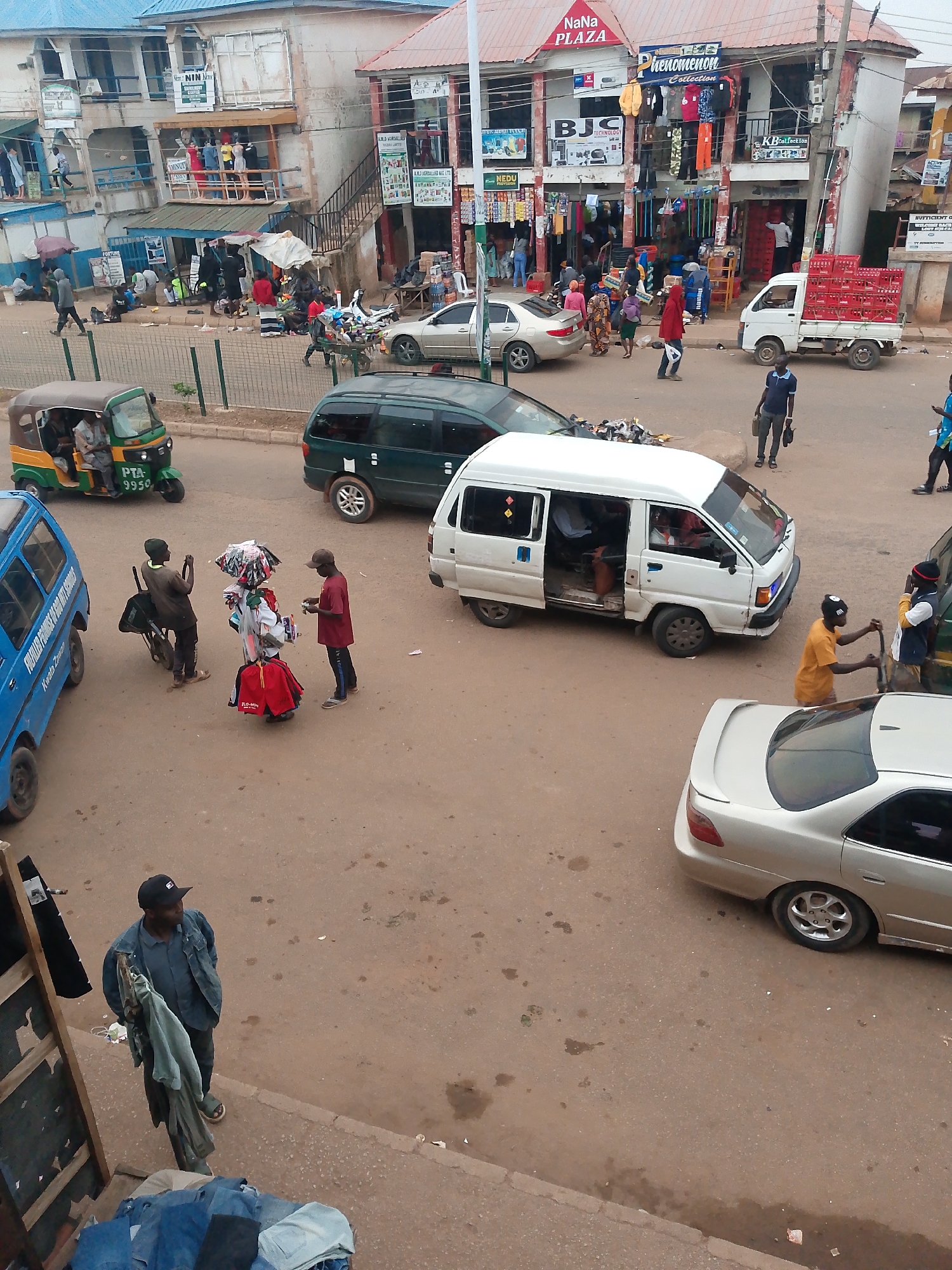 The image captures a street scene in Bukuru, Nigeria, with a focus on a busy commercial area. In the foreground, a man wearing a blue jacket and a cap is standing next to a wooden structure displaying clothes. He is holding several items of clothing. To his right, a silver sedan car is parked on the street.
Further down the street, a white minivan with its doors open is parked. Several people are visible inside and around the minivan. A man in an orange shirt is standing next to the minivan, holding a long object that resembles a pole or a broom. To the right of the minivan, a white truck is loaded with red crates, likely containing beverages.
In the mid-ground, a green minivan is parked on the road. Across the street, a three-wheeled auto-rickshaw, known as an 'okada' or 'keke napep', is visible with passengers. The buildings in the background appear to be shops and commercial establishments, with signage indicating various businesses such as "NaNa PLAZA" and "Phenomenon Collection." People are seen walking on the sidewalks and interacting with street vendors, who are displaying their merchandise on carts and the ground. The overall scene depicts a vibrant and active marketplace.