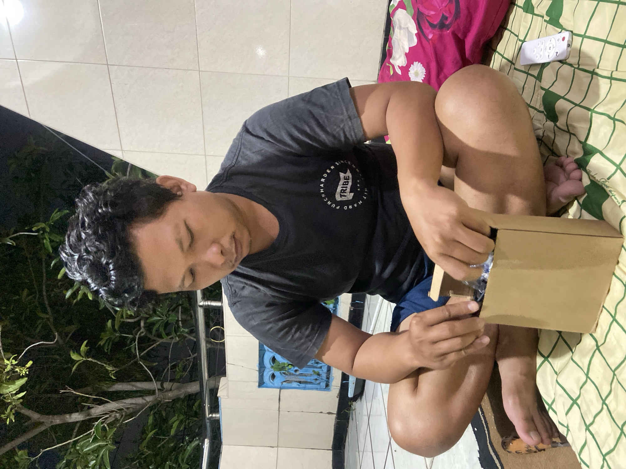 A man squats on a bed or mat in what appears to be an outdoor setting. He is opening a cardboard box. The man is wearing a black t-shirt and blue shorts. A television remote control lies next to him. A pillow with a floral pattern is behind him. The setting seems to be near a building and a tree. The location is Gelam, Indonesia.