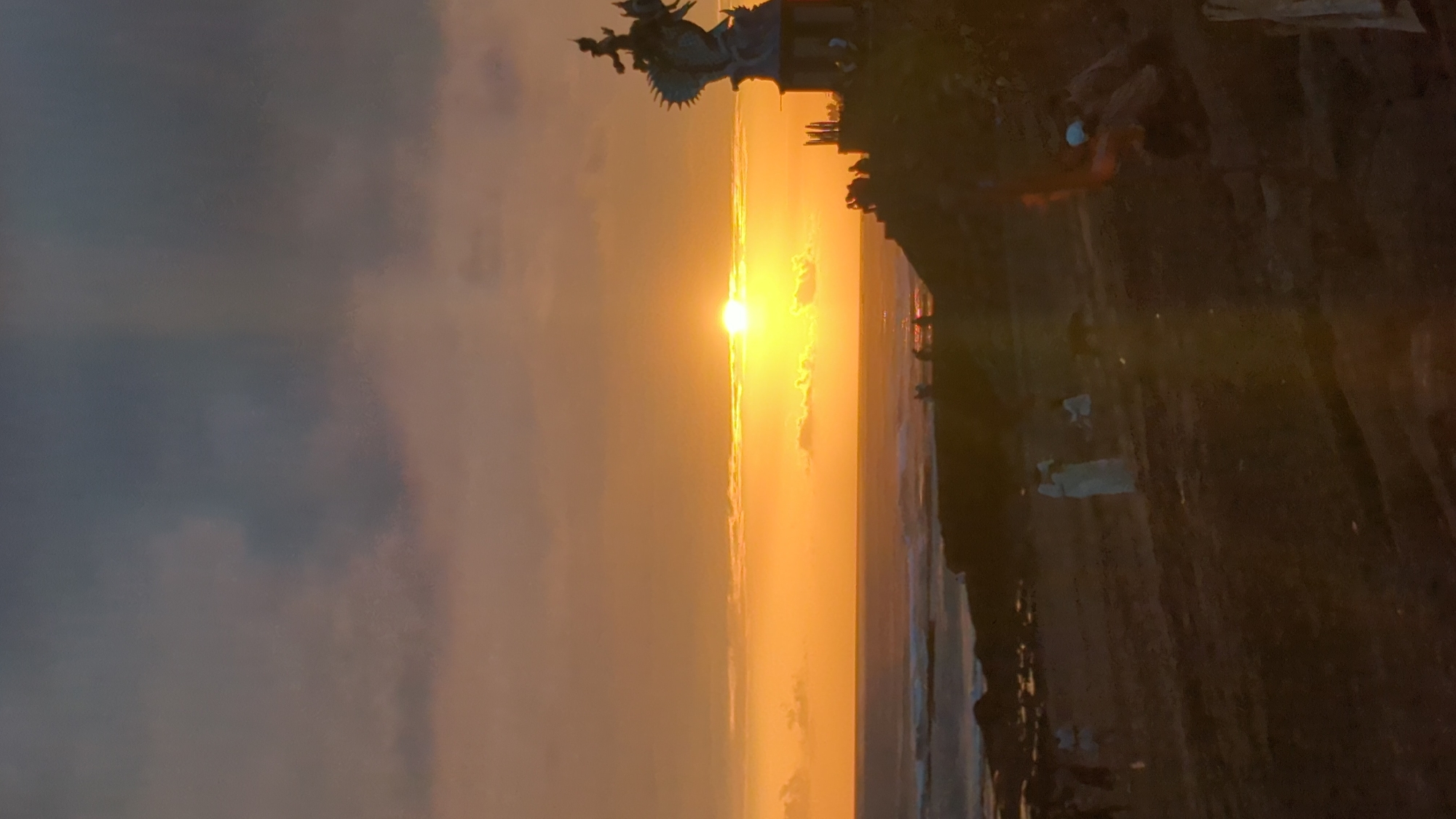 The image depicts a sunset scene at Tanah Lot Temple, located in the village of Beraban, within the district of Kediri, Tabanan Regency, near Mengwi, Indonesia. The sun is setting over the ocean, casting a warm orange glow across the sky and reflecting on the water. In the foreground, a silhouetted statue, likely depicting a mythical creature or deity, stands on a rocky outcrop. Several figures, also silhouetted, are scattered across the beach and the elevated land, some appearing to be tourists or worshippers. Waves are visible crashing against the shore. The overall atmosphere is serene and picturesque, capturing the natural beauty of the location at dusk.
