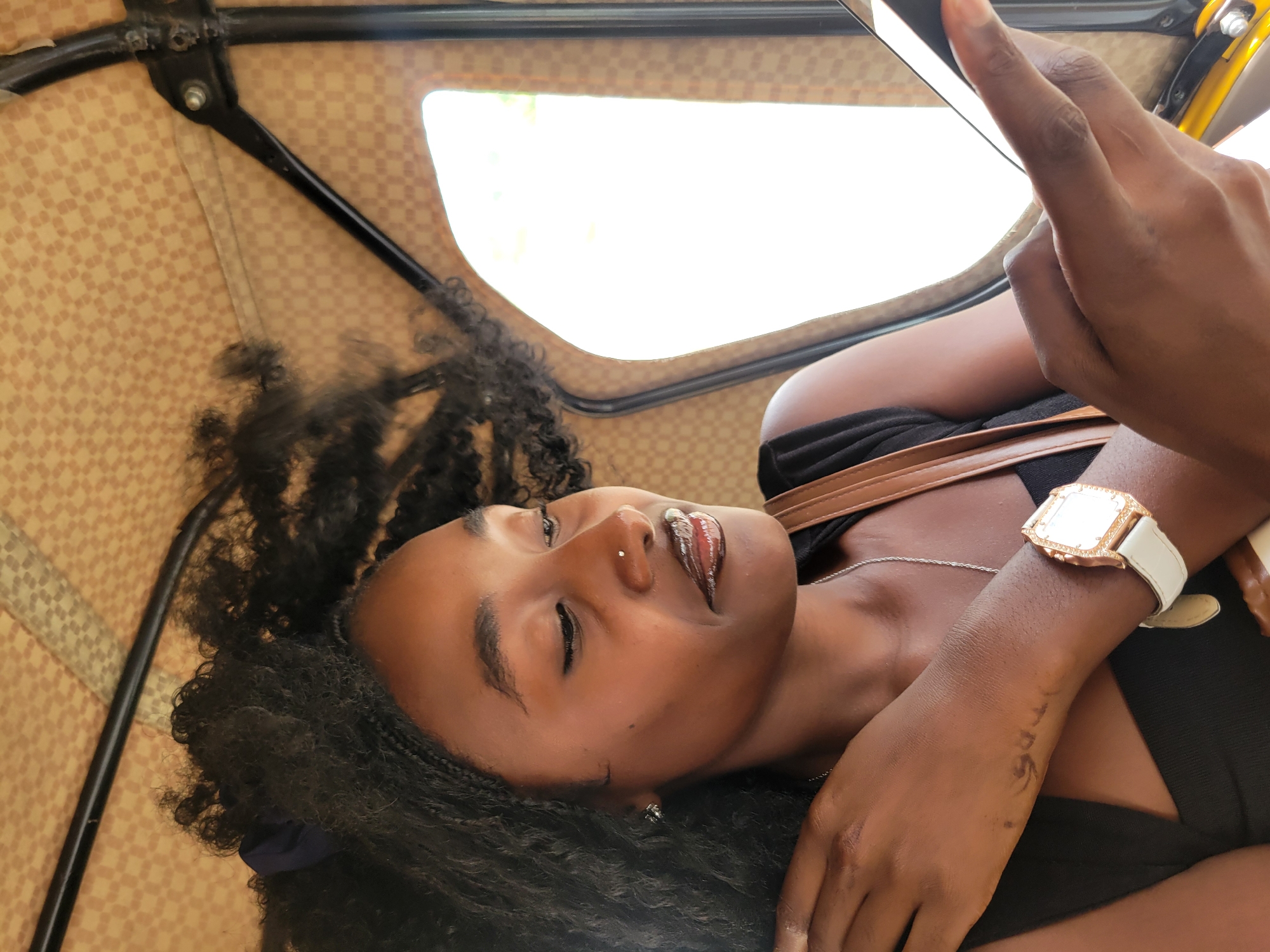 A woman is in the back of a commercial vehicle, likely a tricycle, in Jos, Nigeria. The vehicle's interior has a checkered pattern. The woman is holding a mobile phone. She is wearing a black top and a gold watch with a white band. She has dark, curly hair and a nose piercing. She has writing on her hand.