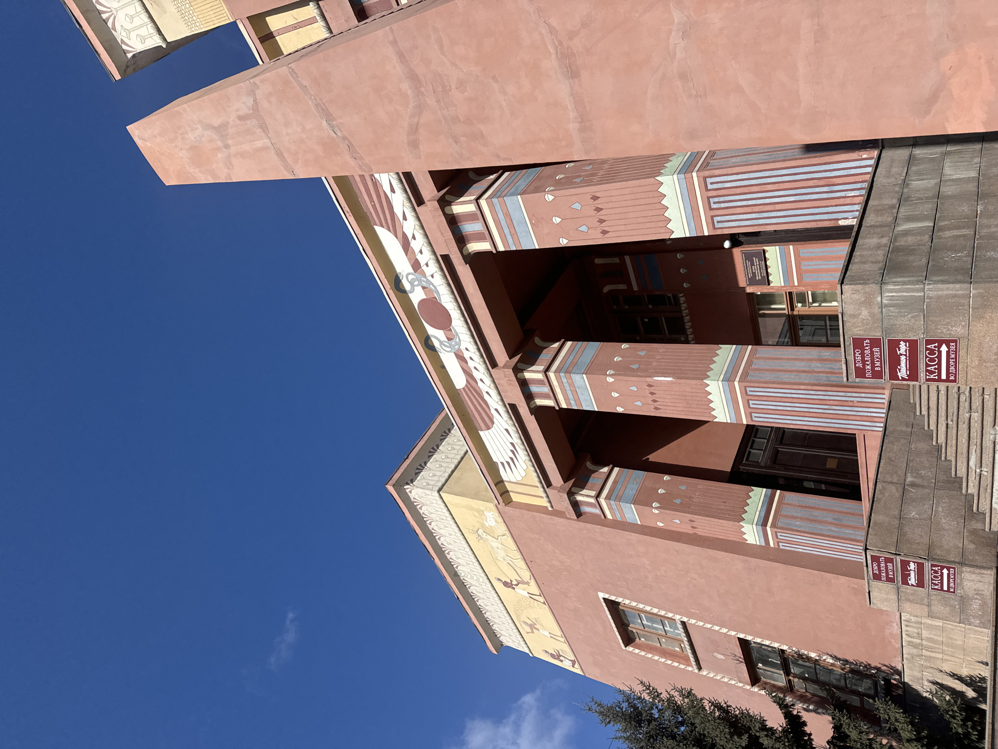 The image depicts a section of the Krasnoyarsk Regional Museum, located in Krasnoyarsk, Russia. The view is from a low angle looking up at the building. The architecture is characterized by a salmon-pink hue and decorative motifs, including stylized imagery of indigenous themes such as stylized animal figures and geometric patterns in colors of brown, white, yellow, blue, and green. Several sets of windows are visible on the side of the building and large rectangular columns leading to a doorway. Various signs are visible, written in Russian, marking the ticket office. A piece of a evergreen tree is visible in the bottom right corner. The sky in the background is blue and free from clouds.