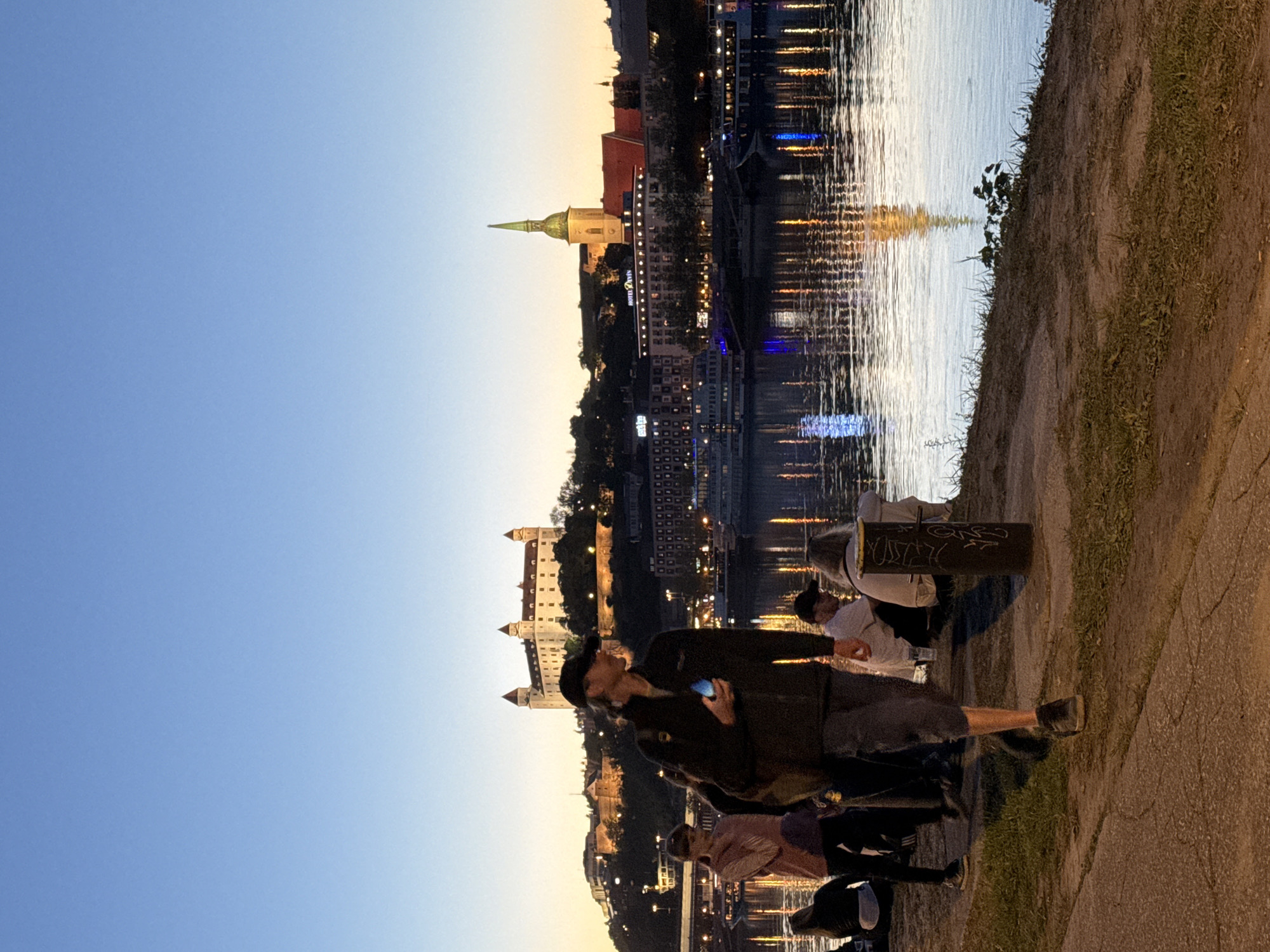 The image shows a waterfront scene in Bratislava, Slovakia, at dusk. A group of people stand along the paved embankment of the Danube River, with the Bratislava Castle and St. Martin's Cathedral prominently visible in the background. Reflections of city lights shimmer on the water's surface. In the foreground, people are looking at their mobile phones and interacting with each other. The sky transitions from blue to orange near the horizon. A black metal post is near the right edge of the embankment. The scene suggests a relaxed, casual atmosphere.
