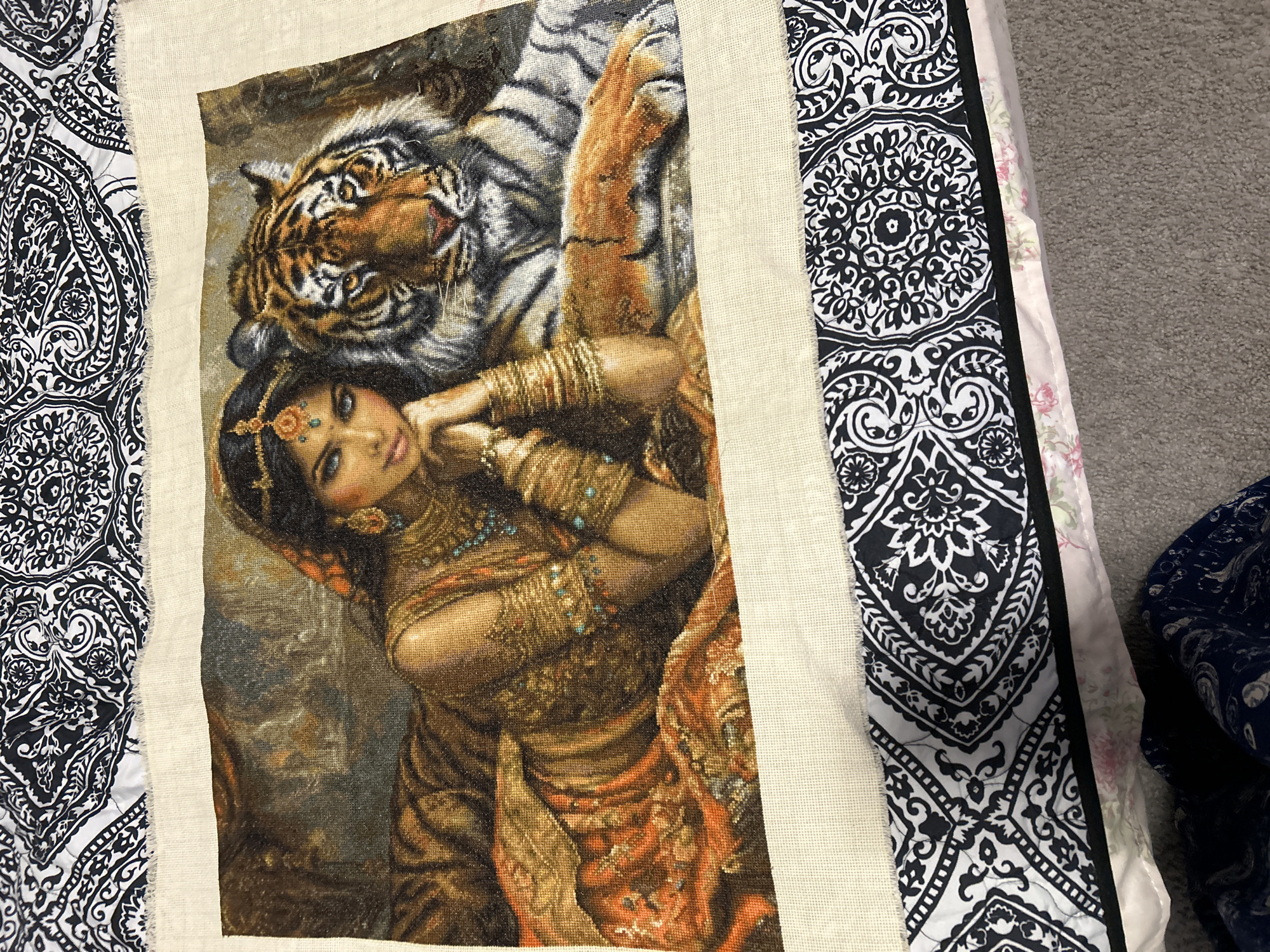 A cross-stitch embroidery depicts a woman with dark hair adorned with gold jewelry and a jewel on her forehead, lying next to a tiger. The woman wears a golden, orange, and brown patterned garment. The tiger is positioned to her right, with its head resting on its paws. The embroidery is laid on top of a cream-colored fabric with frayed edges, which in turn is placed on a surface covered by a black and white patterned fabric. The scene appears to be indoors, possibly in a domestic setting given the bedding underneath. There are no identifiable landmarks or contextual clues to suggest a specific location within Colorado Springs, United States of America.