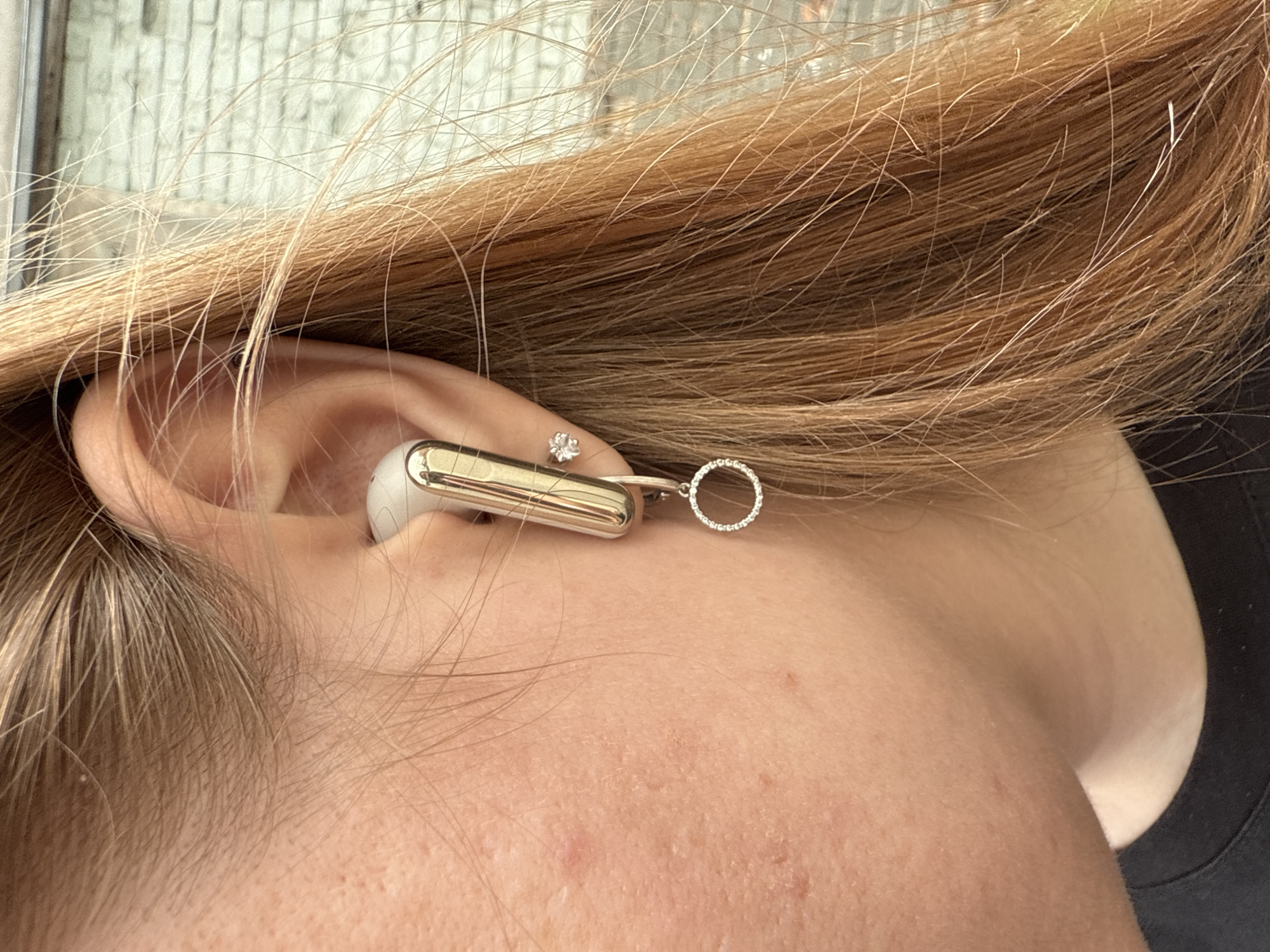 The image is a close-up shot of a person's ear and adjacent skin, with blonde hair framing the ear. A silver earbud with a minimalist design is partially inserted into the ear. Attached to the earbud is a delicate dangling earring featuring a small, sparkling circular charm. A small diamond stud earring is also visible on the earlobe. The lighting suggests it could be daytime, though no specific time of day can be determined. The background is blurred, hinting at an indoor or outdoor setting, but offers no definitive clues about the location beyond what is provided as context. The overall impression is one of personal adornment and technology. There is no visible text in the image.