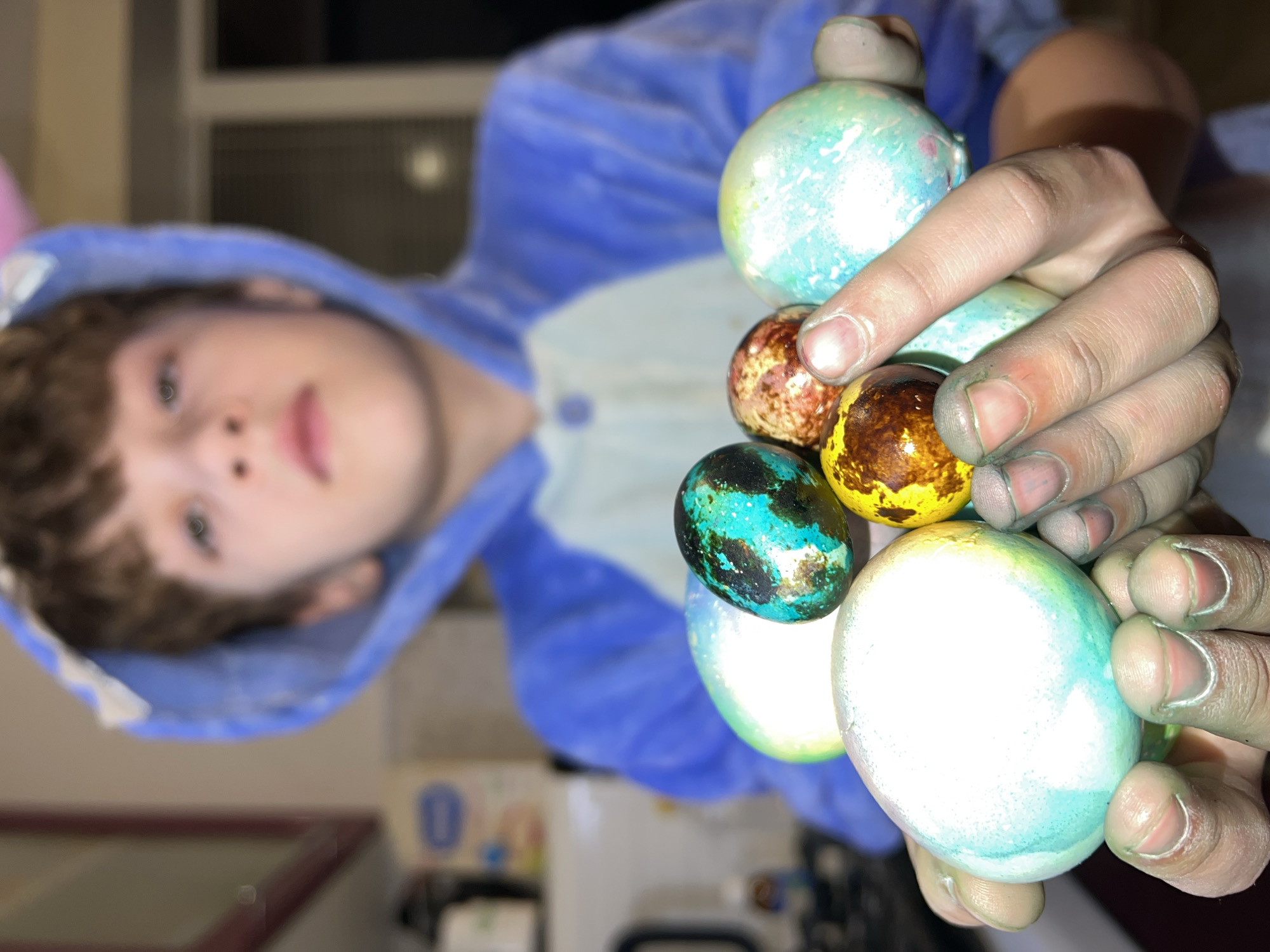 A child wearing a blue hooded onesie with bunny ears holds a handful of brightly colored, decorated Easter eggs in their hands. The child's face is slightly blurred in the background. The eggs feature various patterns and colors, including blue, green, brown, and gold. The background is out of focus but appears to be indoors. The photo is taken at an angle, with the eggs prominently displayed in the foreground. There is no indication of the location of Noviy, Russia in the image.