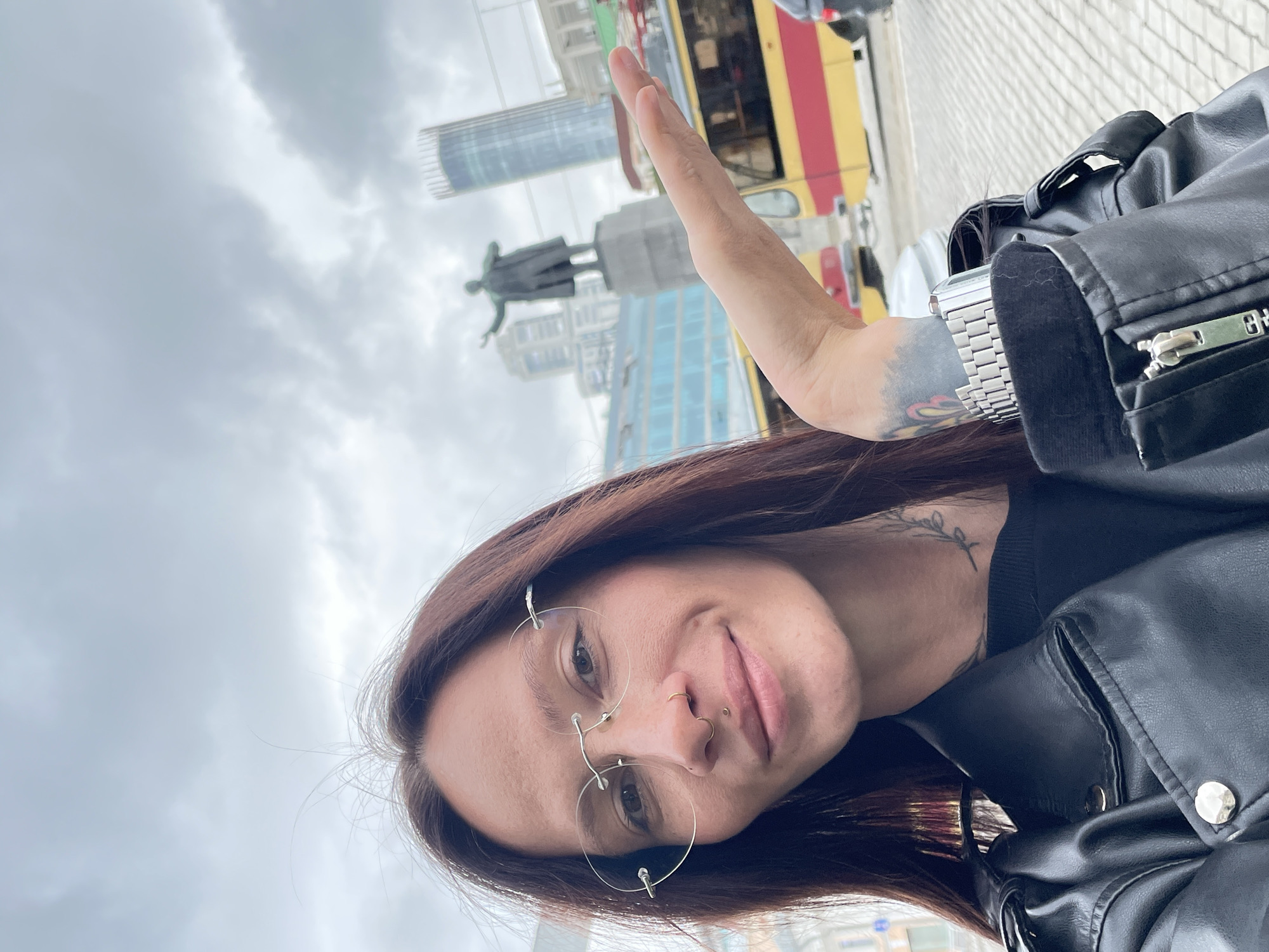 This selfie-style portrait features one individual, a woman with long, dark brown hair, wearing rimless round glasses, a small gold ring in her right nostril, and a philtrum piercing. She wears a black leather jacket over a black top, with a delicate vine tattoo visible on the left side of her neck and a colorful, abstract tattoo on her left inner wrist, adorned with a silver-toned metal link watch. Her right hand is extended with the palm open and slightly cupped, gesturing towards the background.
The background reveals an urban setting under an overcast sky. A prominent bronze statue of Vladimir Lenin stands on a grey stone pedestal, centrally located behind the woman's arm, with his right arm outstretched. Behind the statue and to its right, the tall, cylindrical, glass-clad Vysotsky Business Center skyscraper is clearly visible. To the right of the scene, a yellow and red tram (or trolleybus) is partially visible on the street.
The presence of the Lenin statue and the distinct Vysotsky Business Center definitively places the scene in Yekaterinburg, Russia, specifically near Ploshchad 1905 Goda (1905 Square).
