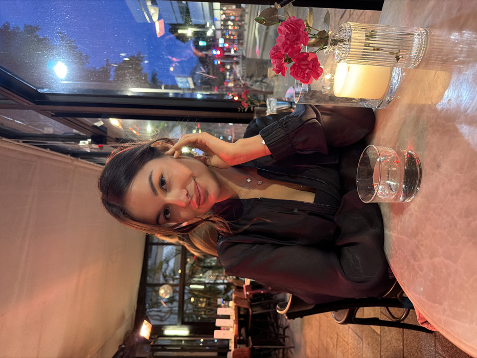 A female individual, appearing to be in her mid-20s to early 30s, is seated at a light-colored table within an indoor dining establishment. She has dark brown hair, parted on the side, and is looking directly at the camera with a subtle smile. She is wearing a dark, possibly black, long-sleeved sheer top over a darker undergarment, along with a silver-toned necklace featuring two distinct pendants and a matching bracelet on her left wrist. Her right hand is positioned near her chin/cheek.
On the table in front of her, a clear glass contains water. To her right, a larger clear cylindrical glass vase holds a lit candle, emitting a warm glow, and next to it, a smaller clear fluted glass vase contains three red artificial flowers.
The interior environment is characterized by warm, artificial lighting, casting an amber/orange hue on the ceiling and elements in the background. Other dark-colored dining chairs and tables are visible further into the establishment. An overhead heater is suspended from the ceiling in the left background.
Through large glass windows behind the individual, an urban street scene is visible at dusk or night. The sky outside is dark blue. Numerous streetlights are illuminated, and red and green traffic lights are discernible in the distance, indicating a busy city thoroughfare. Building silhouettes and trees are visible against the evening sky. The specific location within Tel-Aviv, Israel, cannot be definitively identified from the visible details, but it depicts an urban street scene from within a restaurant or cafe.