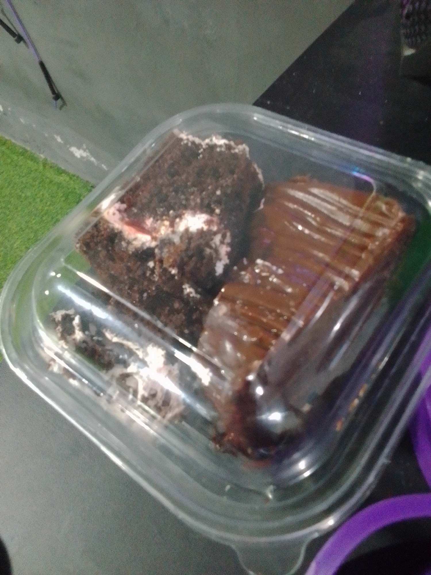 Two slices of chocolate cake are presented in a clear plastic container. One slice is layered with white frosting and appears to have a red filling, while the other slice is fully covered in a dark chocolate glaze. The container is placed on a dark, textured surface, with a patch of artificial green turf visible in the upper left corner. The scene is dimly lit, with reflections from the plastic container adding highlights to the cakes and the surrounding surfaces. The overall impression is that of a pre-packaged dessert. The location is identified as Juan Griego, Venezuela.