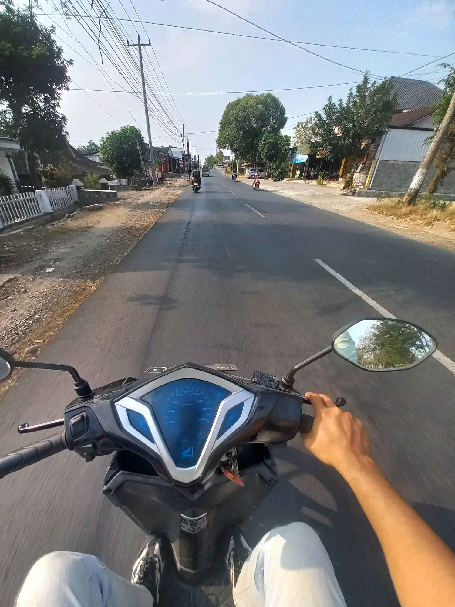 The image is taken from the perspective of someone riding a motorcycle on a paved road in Baturetno, Indonesia. The road is lined with houses, shops, and trees on both sides. Overhead power lines are visible against a clear sky. In the foreground, the motorcycle's dashboard and handlebars are in focus, along with the rider's left leg and hand. The rider's hand is on the throttle, indicating the motorcycle is in motion. A few other motorcycles and a car are visible further down the road. The general scene suggests a typical day in a rural or semi-urban Indonesian setting.