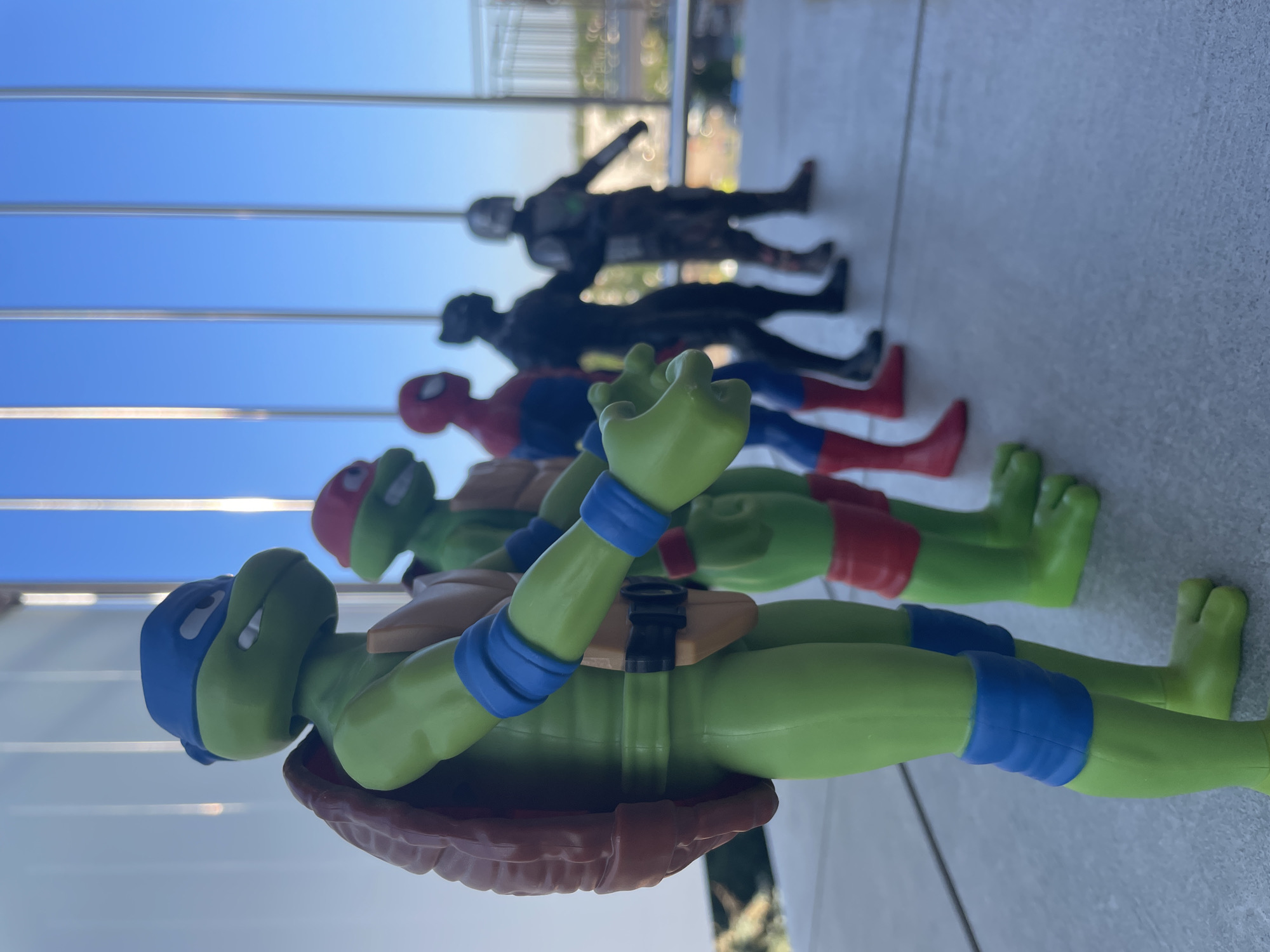In an outdoor urban setting, a line of action figures stands on a concrete ledge against a backdrop of a clear blue sky and distant city buildings seen through vertical architectural elements. The figures, arranged from left to right, are Leonardo, Michelangelo, Spider-Man, Black Panther, and The Mandalorian. Leonardo, the leftmost figure, is predominantly green with a blue mask, brown shell, and orange chest harness with blue armbands and knee pads. Michelangelo is also green with a red mask, orange chest harness, and red armbands and knee pads. Spider-Man is in his iconic red and blue suit. Black Panther is a silhouetted black figure. The Mandalorian figure is dark grey with a lighter grey chest plate and red accents on its legs. The figures are facing to the right, with Leonardo's hands open and extended forward. The focus is on the figures, with the background slightly blurred.