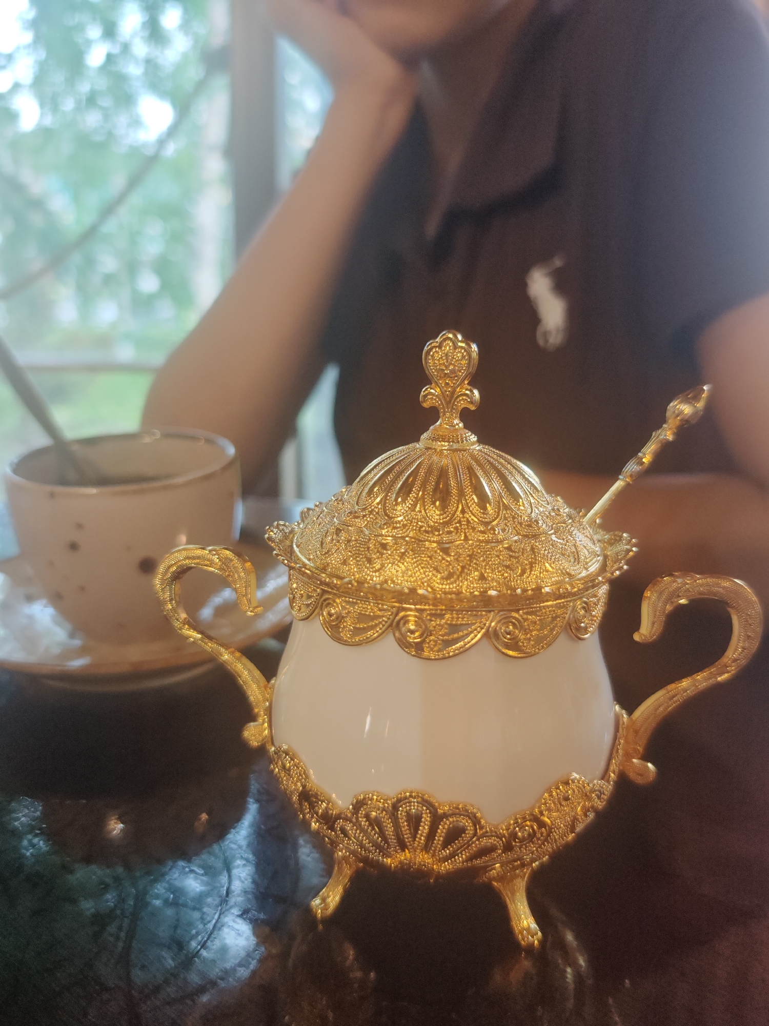 The image shows a close-up of an ornate, white ceramic sugar bowl with gold-colored filigree accents, a decorative lid, and a matching spoon. The sugar bowl is placed on a dark, reflective surface, possibly a table. To the left of the sugar bowl is a speckled ceramic teacup with a spoon resting inside, and a saucer. In the background, slightly out of focus, a person wearing a dark polo shirt with a white logo is visible, with their arm resting on their head. The setting appears to be indoors, near a window with greenery visible outside, suggesting a daytime scene. The overall impression is one of a relaxed moment, perhaps during a tea or coffee break. There is no visible text in the image.