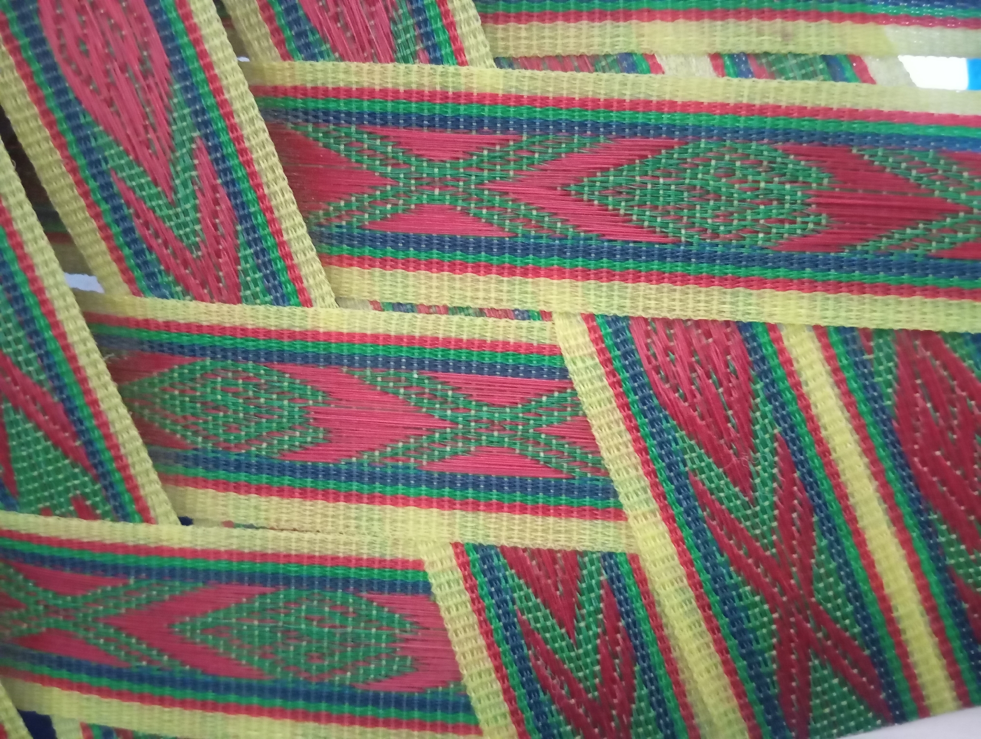 The image is a close-up, top-down view of a woven mat with a colorful geometric pattern. The mat is made of thin, flexible strips of material, predominantly yellow, red, green, and blue. The pattern features horizontal bands of color interspersed with diamond-shaped designs formed by the red and green threads. The weave is tight, creating a textured surface. The mat appears to be layered or folded in some areas, with strips overlapping each other. The lighting is even, with no distinct shadows, suggesting an indoor setting or diffused outdoor light. There are no people or other objects visible in the frame, and the background is indistinct, allowing the focus to remain entirely on the texture and pattern of the mat. The scene is static and devoid of action. The location is unknown.