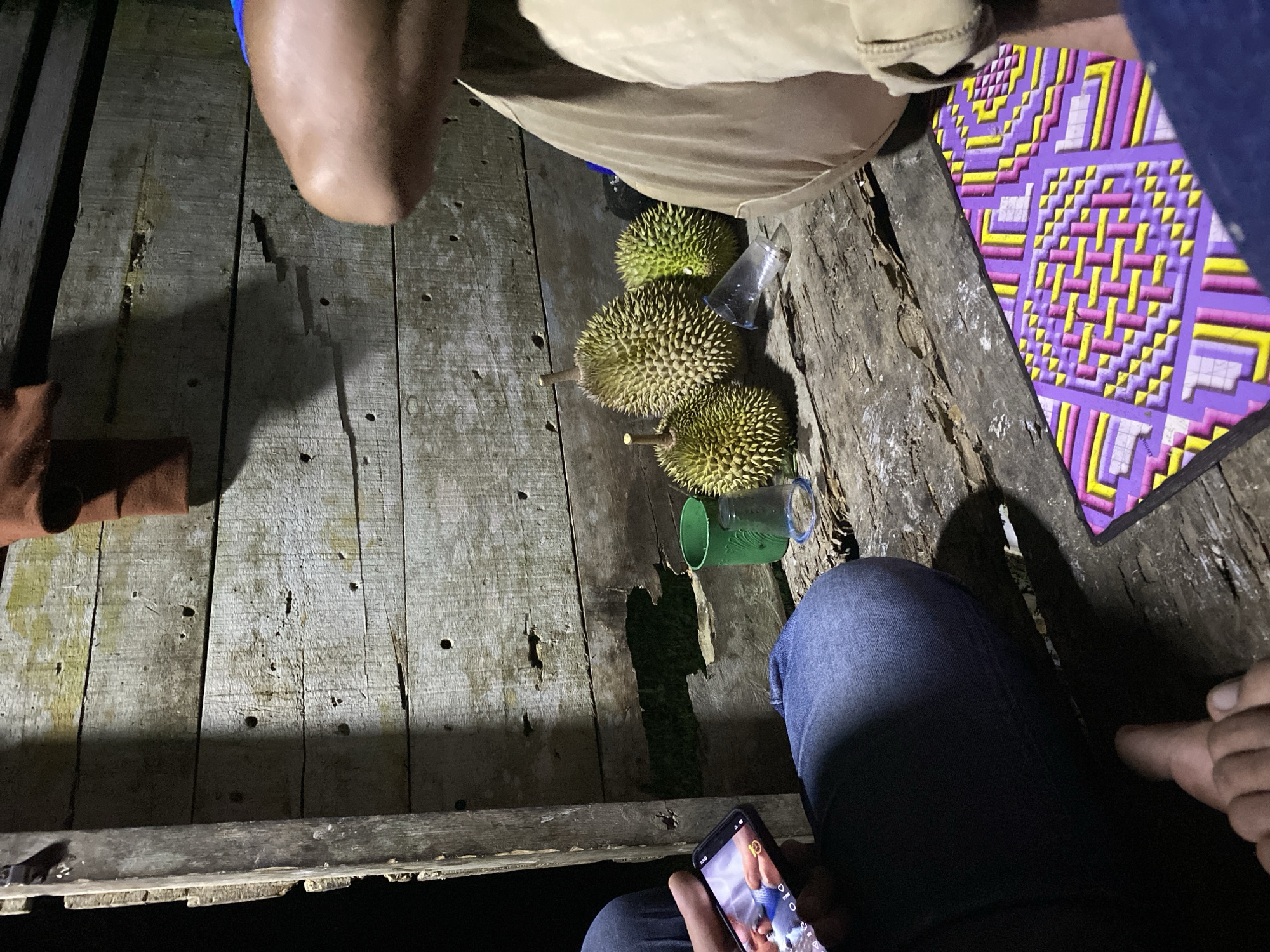 This is a low-light, medium-shot photograph taken outdoors at night, likely in the village of Lamtui, Indonesia, given the context of durian fruit. The scene is set on a rustic wooden surface, possibly a porch or deck, with a weathered wooden wall as the backdrop. In the foreground, a person wearing dark blue jeans is seated on the left, holding a smartphone that displays an image. Their left hand is visible, holding the phone. To the right of this individual, a collection of three durian fruits is arranged on the wooden surface. One durian is in the center, appearing larger and spikier than the two smaller ones flanking it. A green plastic cup and a clear plastic cup lie near the durians. Further to the right, another person is seated, wearing light brown or khaki pants. Their legs and lower torso are visible, and their knee is prominent. The ground is a rough wooden plank. A colorful mat with geometric patterns in purple, yellow, and pink is placed on the wooden surface in the lower right corner of the image, partially beneath the person in khaki pants. The wooden wall behind the subjects shows signs of age and weathering, with visible grain and scattered holes. A dark fabric, possibly clothing, hangs from the top of the wall on the left. The lighting is dim and atmospheric, casting shadows and highlighting the textures of the wood and the durian fruits.
