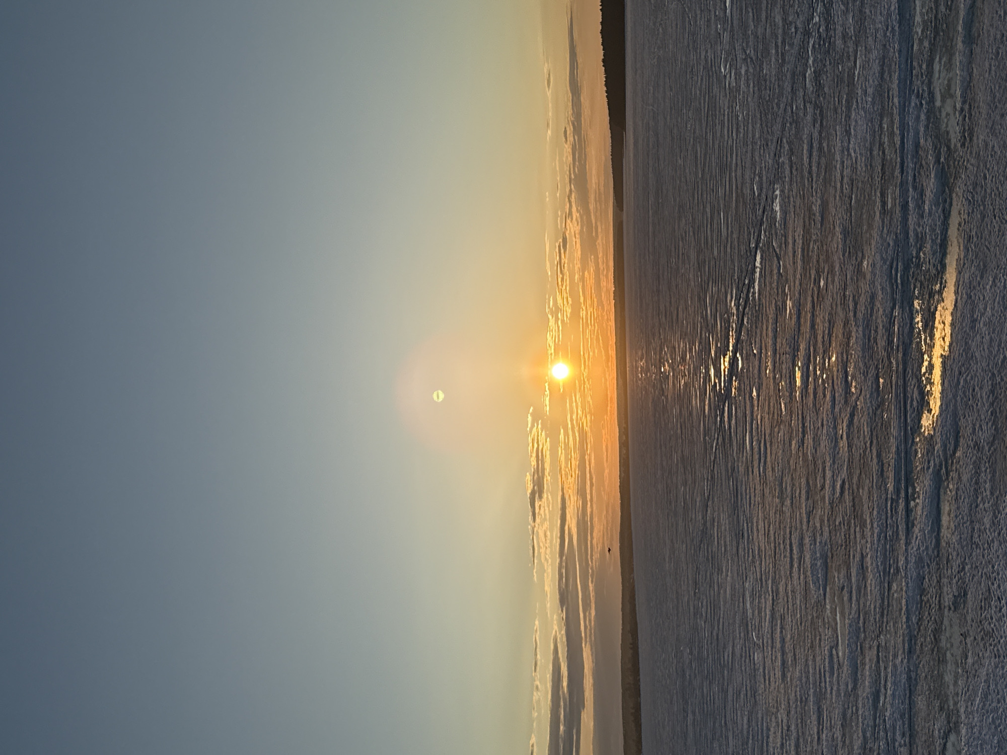 The image shows a sunset over a frozen body of water in Votkinsk, Russia. The sun is positioned on the horizon, casting a bright yellow glow. The sky is a gradient, transitioning from a light blue on the left to a yellow-orange near the horizon. The frozen water reflects the sunlight, appearing textured and icy. A dark line of trees or land is visible along the horizon.