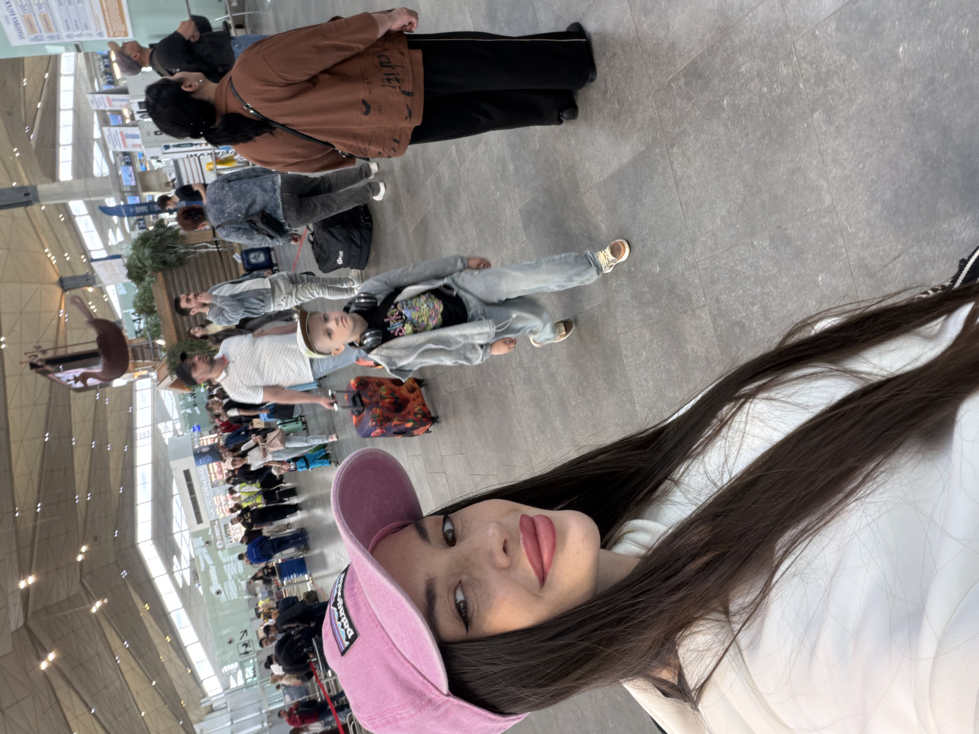 A candid, vertically-oriented image captures a young woman taking a selfie in what appears to be a busy airport or large transport terminal in Saint Petersburg, Russia. The woman, with long dark hair, wears a pink Patagonia baseball cap, a white top, and features bold pink lipstick, looking directly at the camera with a slight smile.
Behind her, numerous people are scattered across a spacious, light-grey tiled floor. In the midground, a child with light hair and headphones around their neck is visible, wearing a colorful graphic t-shirt, light jeans, and a grey hoodie, sitting or lying on the floor. Further back, an adult in a brown jacket and black pants stands with their back to the camera, next to another person in a denim jacket. Many other individuals are seen in the background, some pulling luggage, suggesting they are waiting in queues or moving through the terminal.
The setting is a large indoor public space with a high ceiling featuring prominent structural beams and skylights, indicating a modern architectural design and natural light. Artificial lighting fixtures are also visible. A small potted plant adds a touch of green to the utilitarian environment. The overall activity suggests transit, with people waiting and navigating a bustling area. The time of day appears to be daytime due to the natural light. The only clearly discernible text is the "Patagonia" logo on the woman's cap.