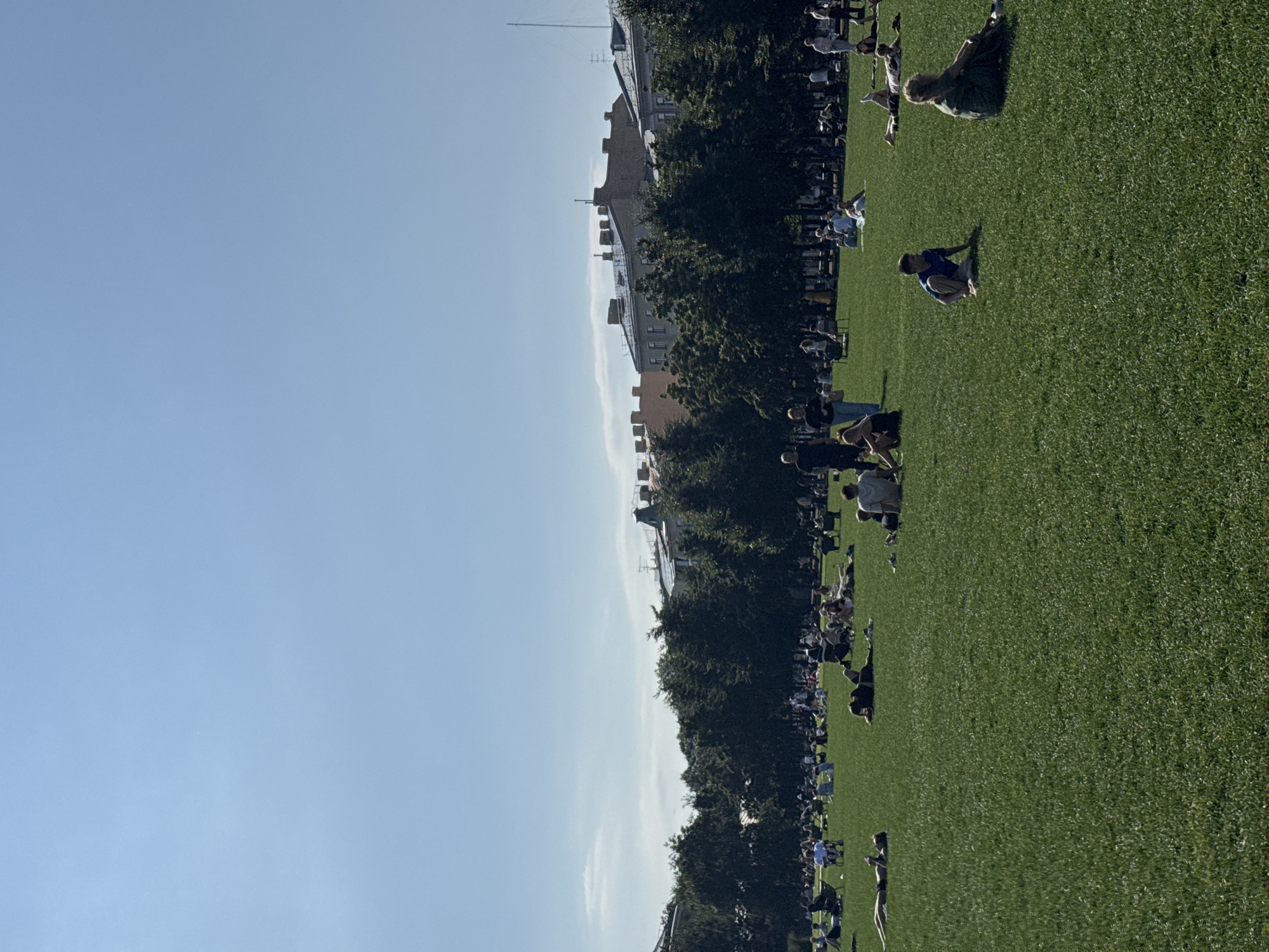 The image shows a group of people spread out on a lush green lawn under a clear blue sky. The perspective is from above, looking down at the scene. Many individuals are engaged in a yoga or exercise session, with some in various poses, while others are simply sitting or lying on the grass. The setting appears to be a park or a large open grassy area, bordered by trees and buildings in the background. The buildings suggest an urban or suburban environment.
The time of day seems to be daytime, likely late morning or afternoon, given the bright sunlight casting shadows. The weather appears to be pleasant and sunny. There are no visible text elements in the image. The overall atmosphere seems relaxed and active, with people enjoying outdoor activities. The individuals are dressed in casual attire suitable for exercise or leisure. The scene suggests a community gathering or a class taking place in an open-air setting. The people are diverse in age and gender. The focus is on the people and their engagement with the activity and the environment.