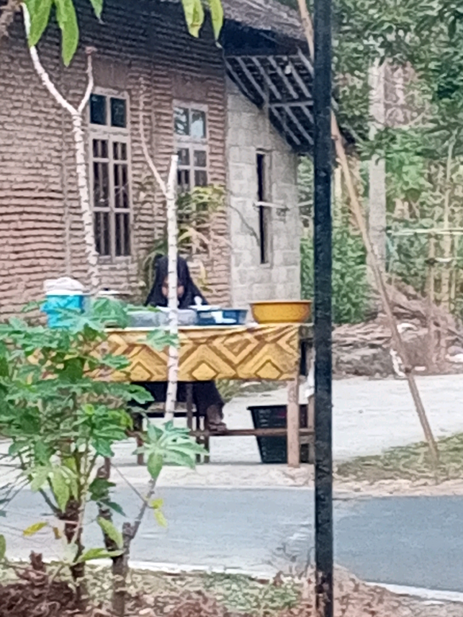 The image depicts an outdoor scene in Kedungringin, Indonesia. In the foreground, lush green foliage obscures the lower portion of the frame. A dark, vertical pole stands prominently in the right half of the image, bisecting the view. Behind the pole, a woman wearing a black head covering is seated at a table. The table is draped with a patterned cloth featuring yellow and brown geometric designs. On the table, there are several objects, including a yellow bowl, a blue container, and what appears to be food or other items. In the background, a brick building with windows is visible, along with part of a roof structure. Trees and vegetation surround the building, suggesting a rural or semi-rural setting. The overall lighting suggests it is daytime.