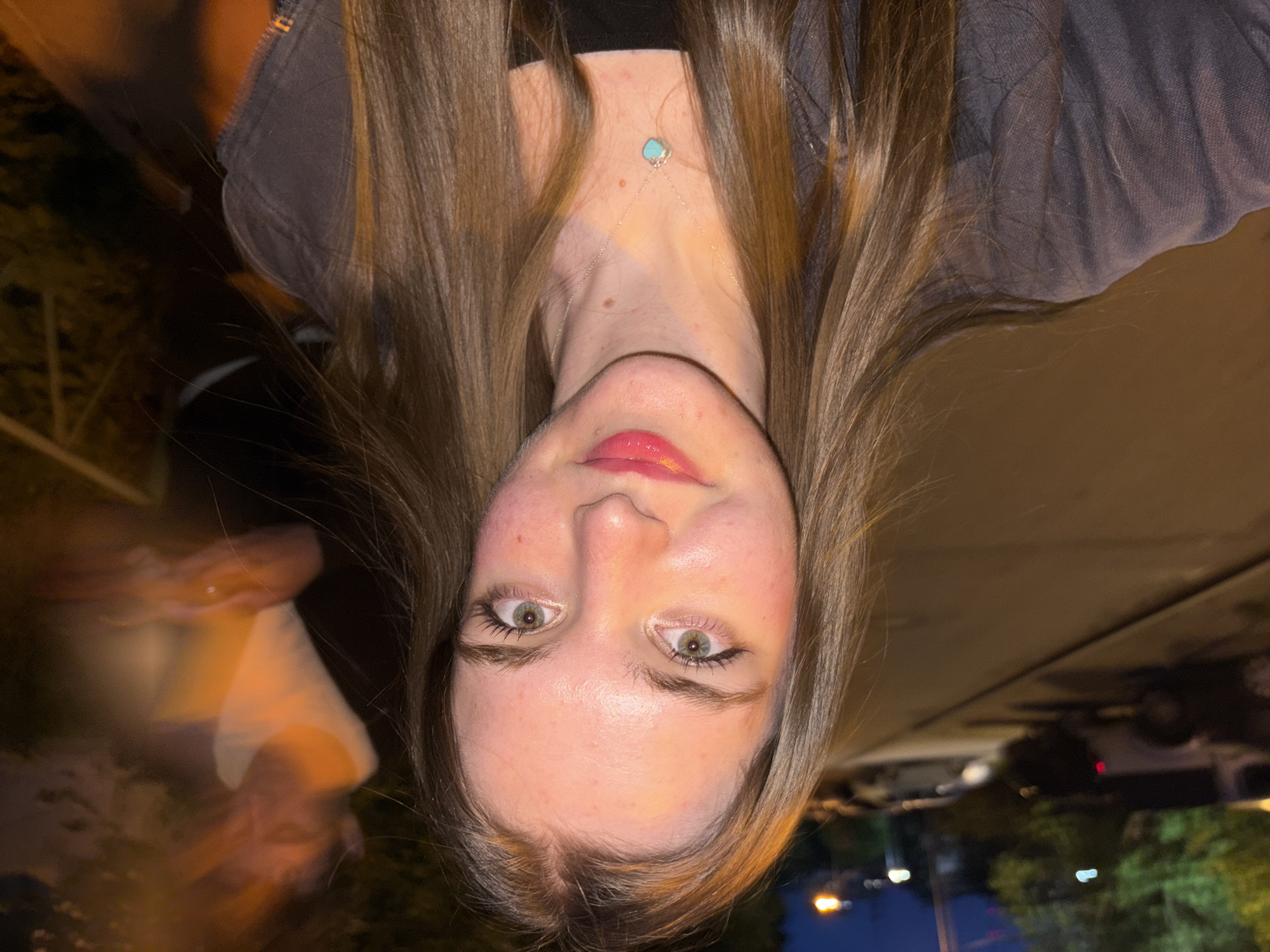 A young woman is the primary subject of this upside-down, close-up photograph, which appears to be taken outdoors. The woman has fair skin, green eyes, and light brown hair that frames her face and extends downwards. She is wearing lip gloss and a necklace with a turquoise stone. She is wearing a dark-colored top underneath a gray jacket. The background is blurred and indistinct. There are blurred lights and trees, as well as what appears to be street pavement and a parked car. There is no evidence in the image that connects it to the city of Perm, Russia. The focus is on the woman's face, and there are no actions or interactions readily apparent in the image.